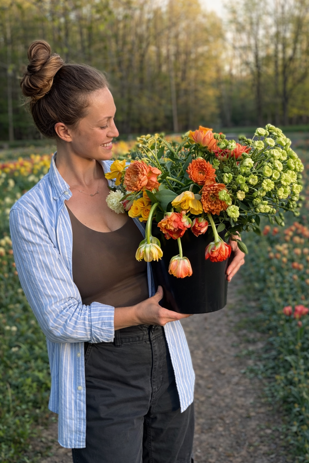 A woman smiling while holding a large pot of colorful flowers in a field of blooming flowers during sunset.