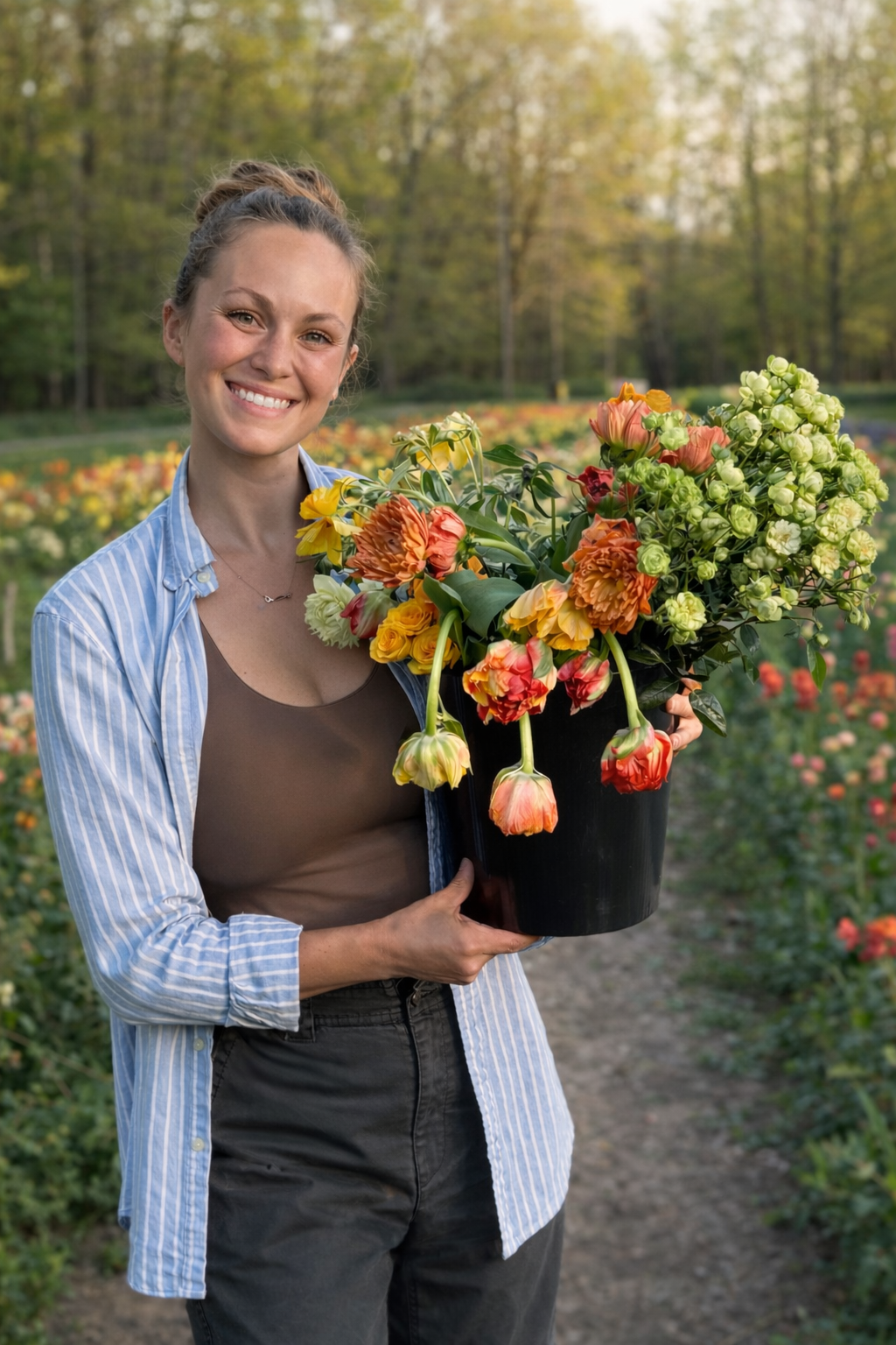 A smiling woman holding a large black pot filled with colorful flowers, standing in a garden of blooming flowers with trees in the background.