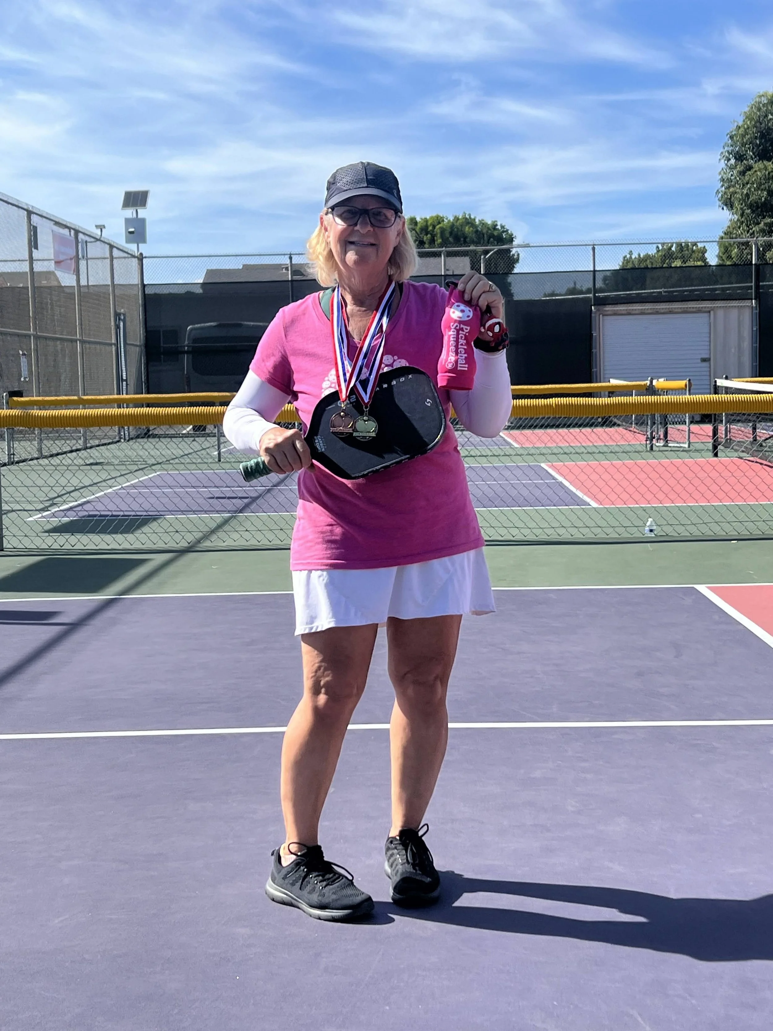 An older woman standing on a tennis court, wearing medals around her neck, holding a tennis paddle, and displaying a pink item and keychain, smiling at the camera.