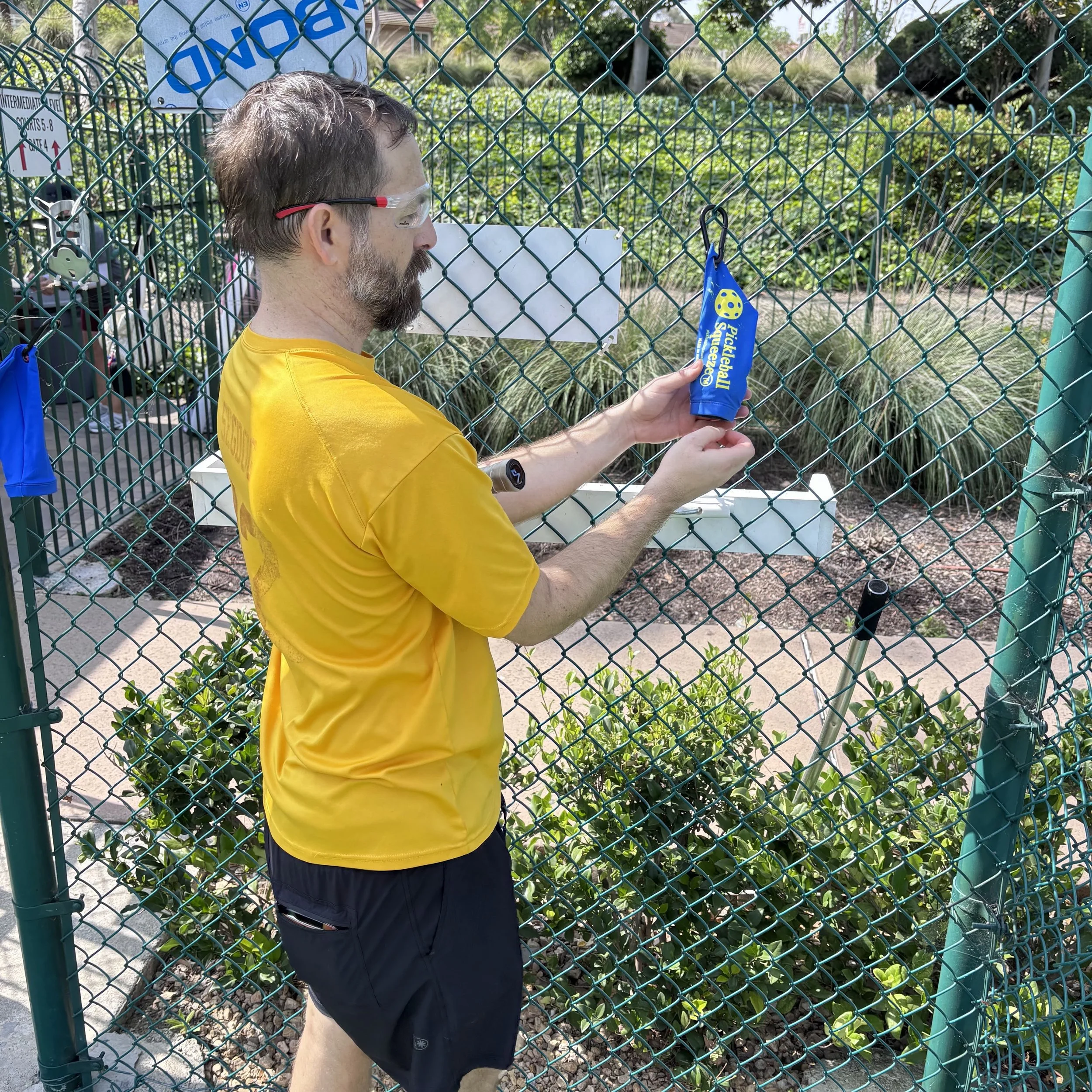 A man in a yellow sports shirt and black shorts stands outside a green chain-link fence, holding a blue tube labeled 'Pickleball' near a pickleball court. The man is wearing safety glasses and a wristwatch, and there are plants and other pickleball e