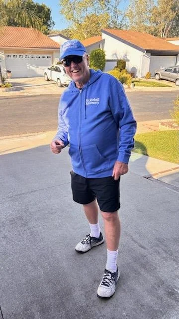 Older person wearing a blue hoodie, black shorts, and athletic shoes, standing outside on a driveway, smiling at the camera.