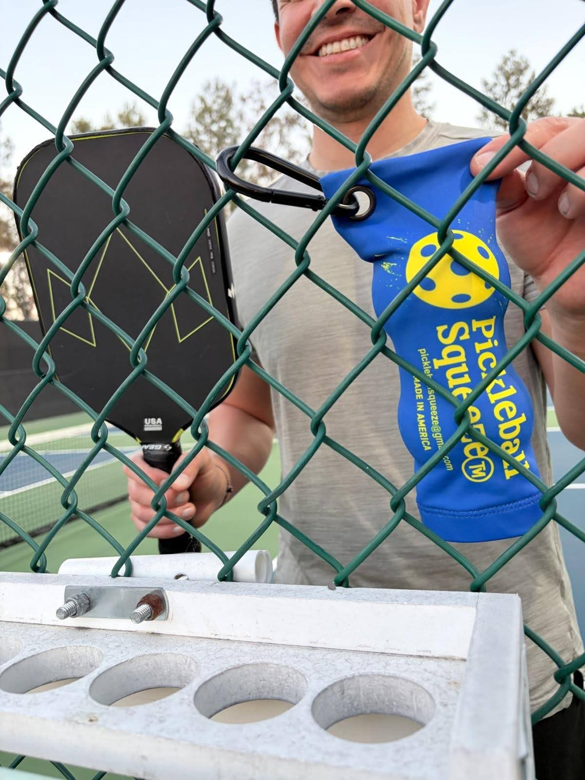 Person smiling behind a chain-link fence on a tennis court, holding a pickleball paddle, and hanging a blue pickleball squuezer with a yellow smiley face logo on the fence.