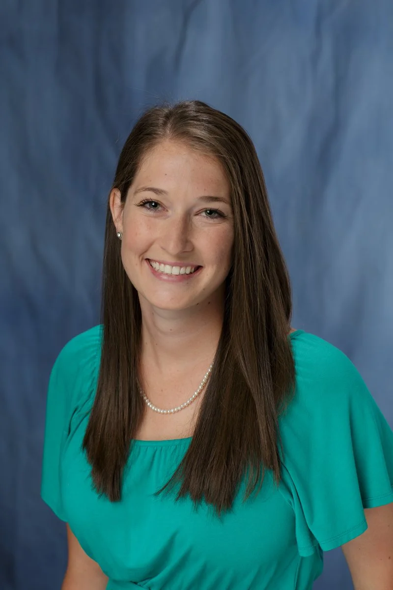 A smiling woman with long brown hair, wearing a turquoise blouse and a pearl necklace, standing against a blue backdrop.