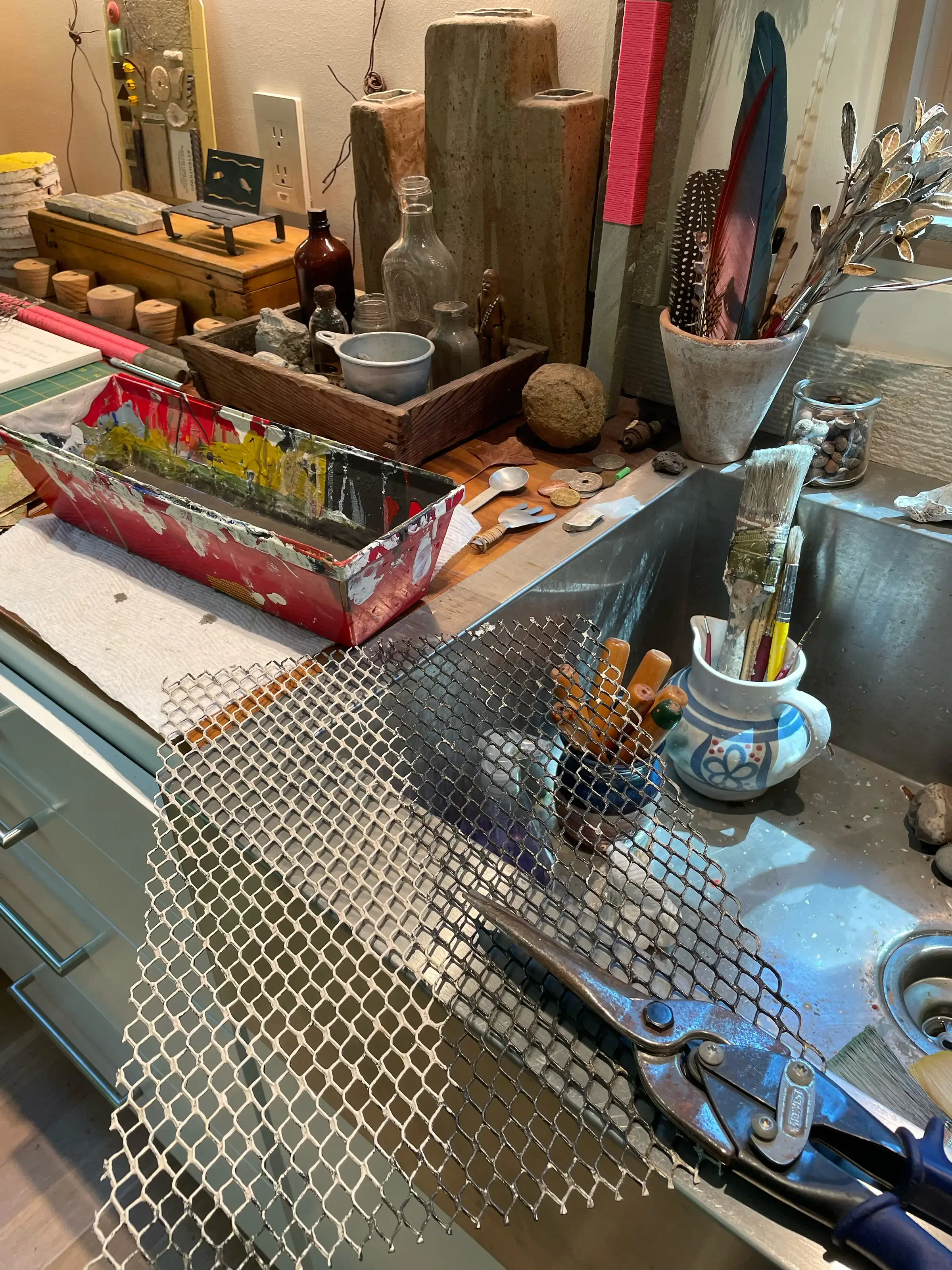 A cluttered artist's workspace and kitchen sink with various art supplies, bottles, and decorative items.
