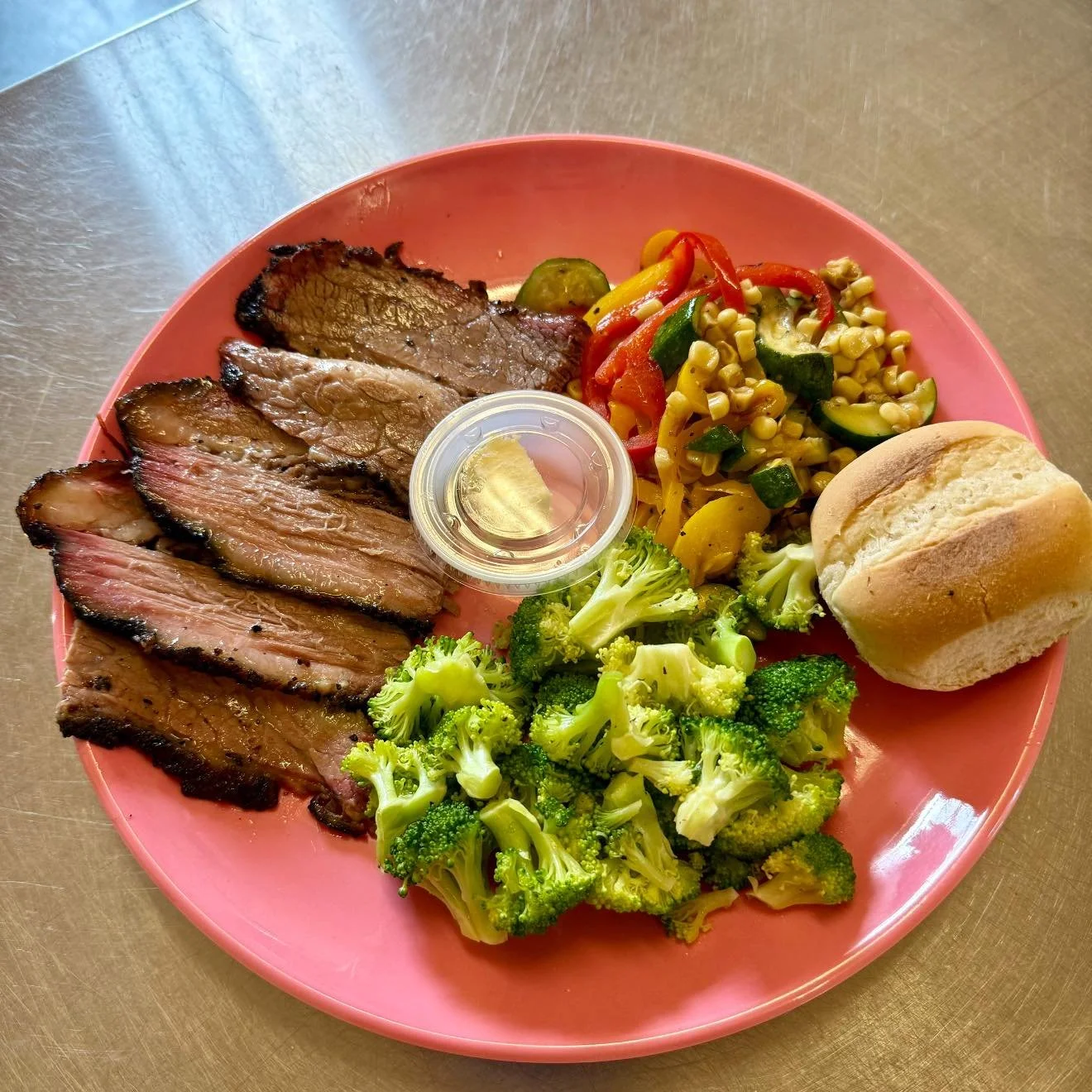 A pink plate with slices of cooked meat, a small cup of clear juice, steamed broccoli, a mixed vegetable salad with corn, cucumbers, and peppers, and a bread roll.