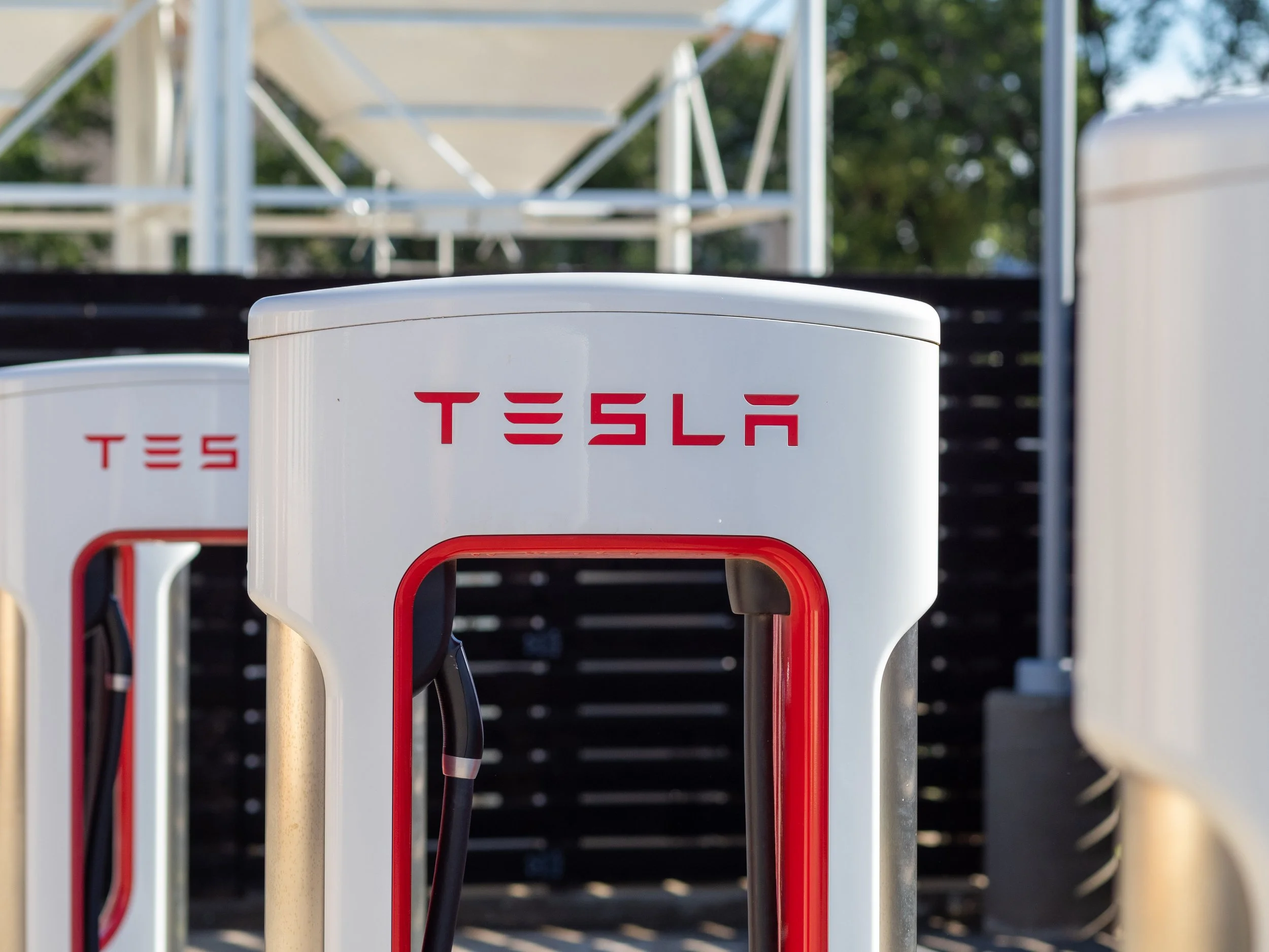 Tesla electric vehicle charging stations outdoors with electrical cables and charging ports, with trees and structures in the background.
