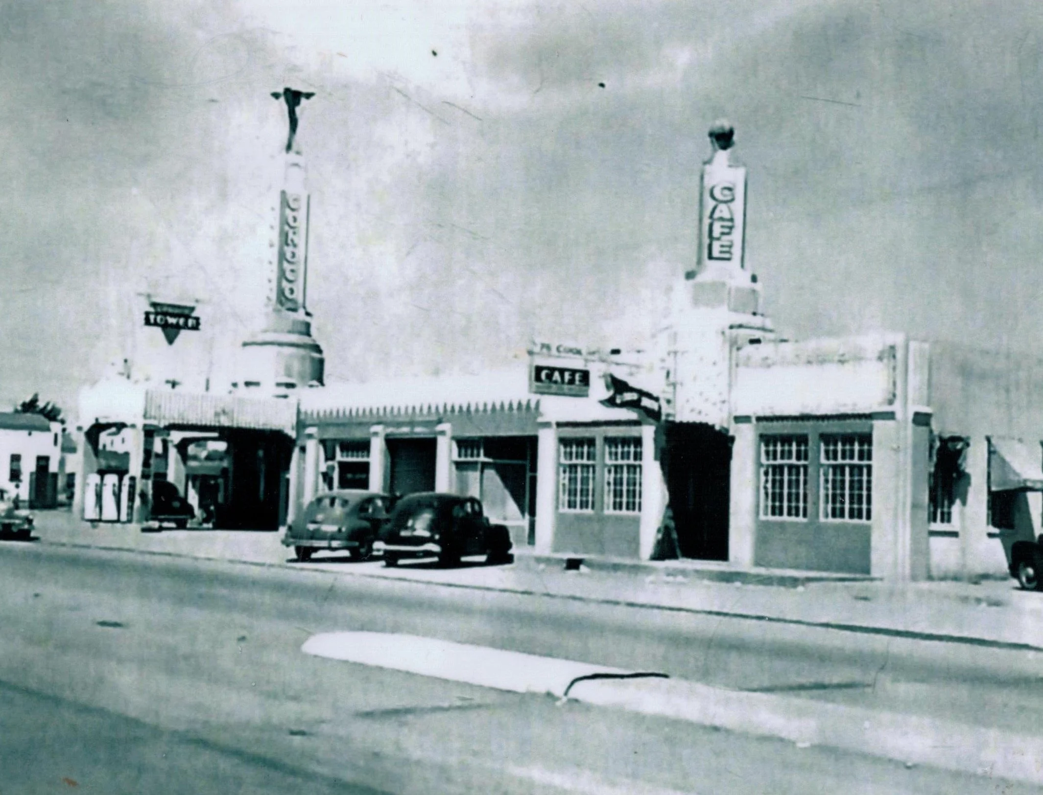 Black and white photograph of a street scene with a large building that has tall columns, signs, and several cars parked in the foreground.