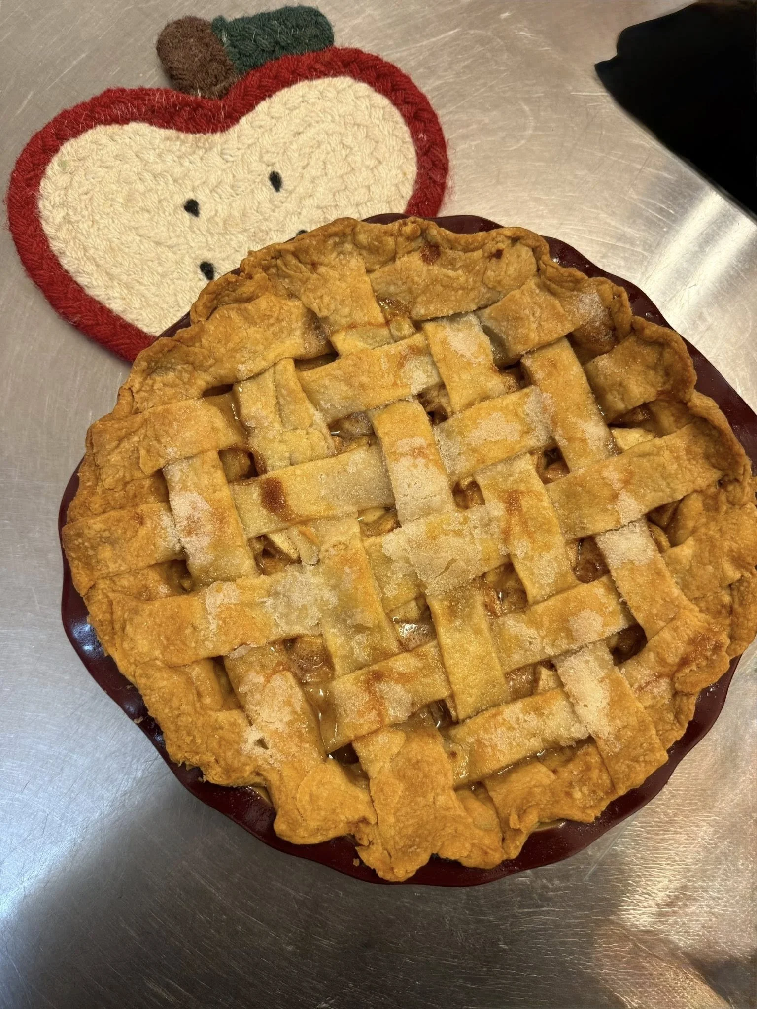 A freshly baked apple pie with a lattice crust on a dark plate. A Hello Kitty-themed heat-resistant mat or cloth with a face and bow is partially visible underneath the plate. The pie is on a metallic surface.