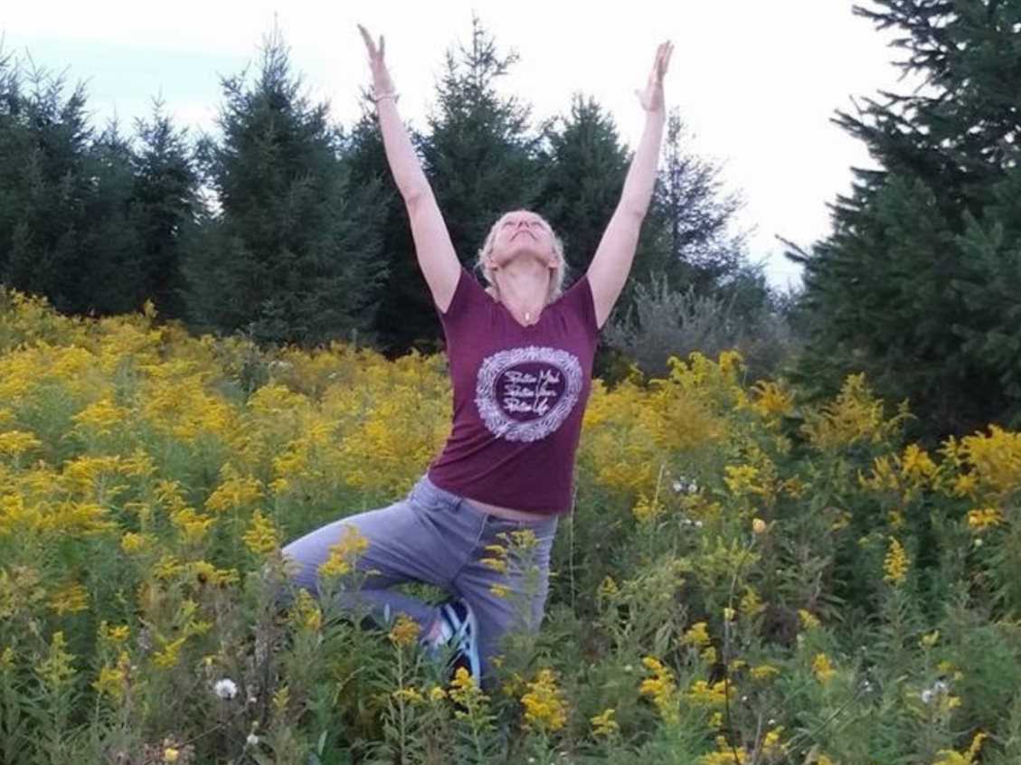 Woman practicing yoga outdoors in a field of yellow flowers with pine trees in the background, arms raised and face upward.