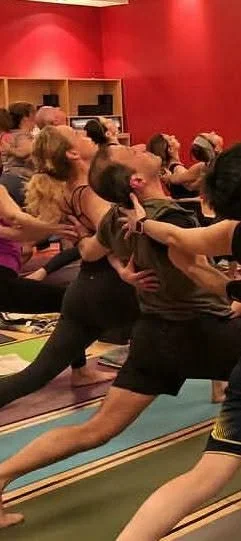 A group of people practicing yoga together in a studio, performing a warrior pose. The studio has red walls and wooden shelves.