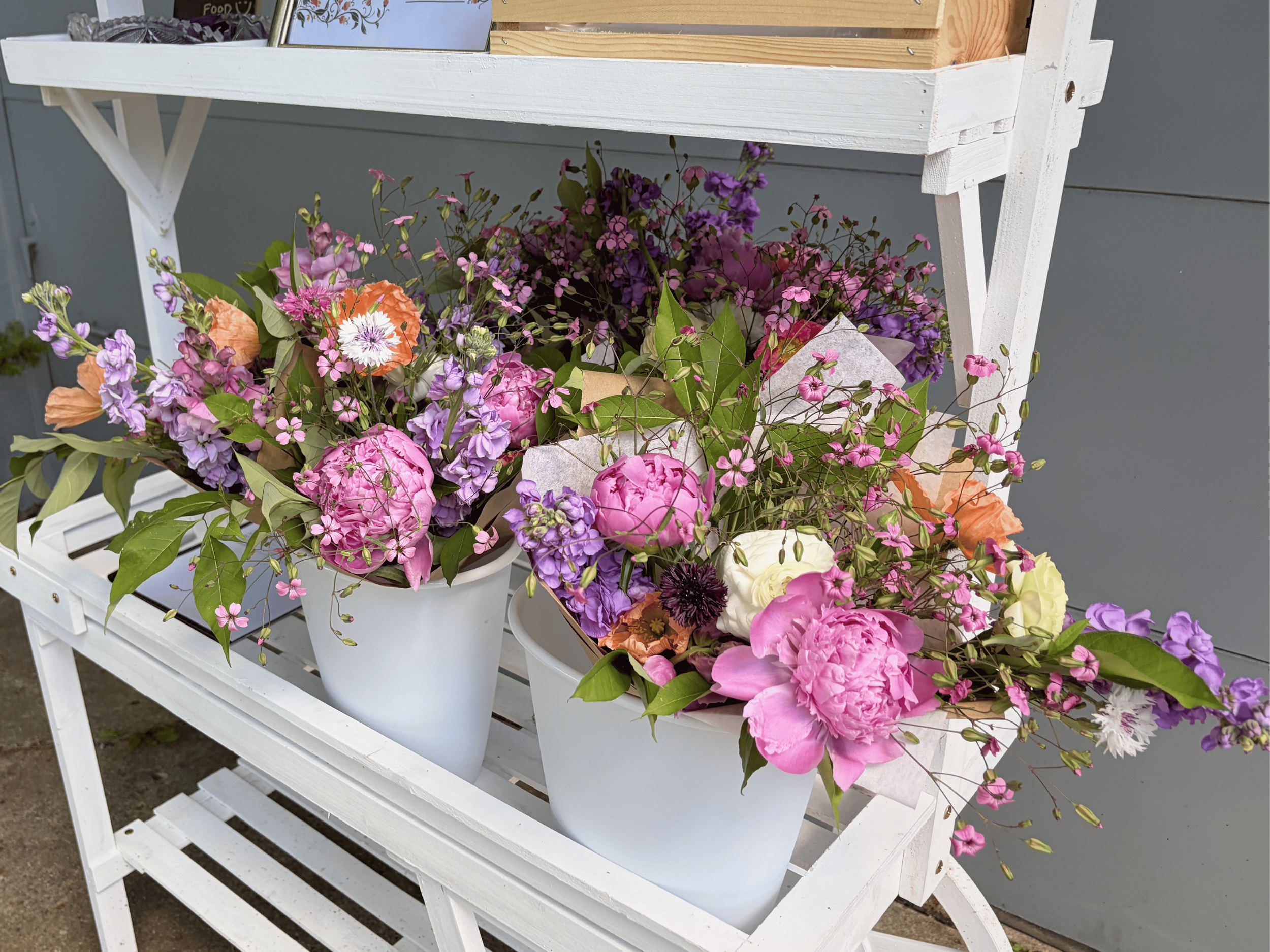 Colorful flower bouquets with pink, purple, orange, and white blossoms arranged in white pots on a white wooden display shelf.