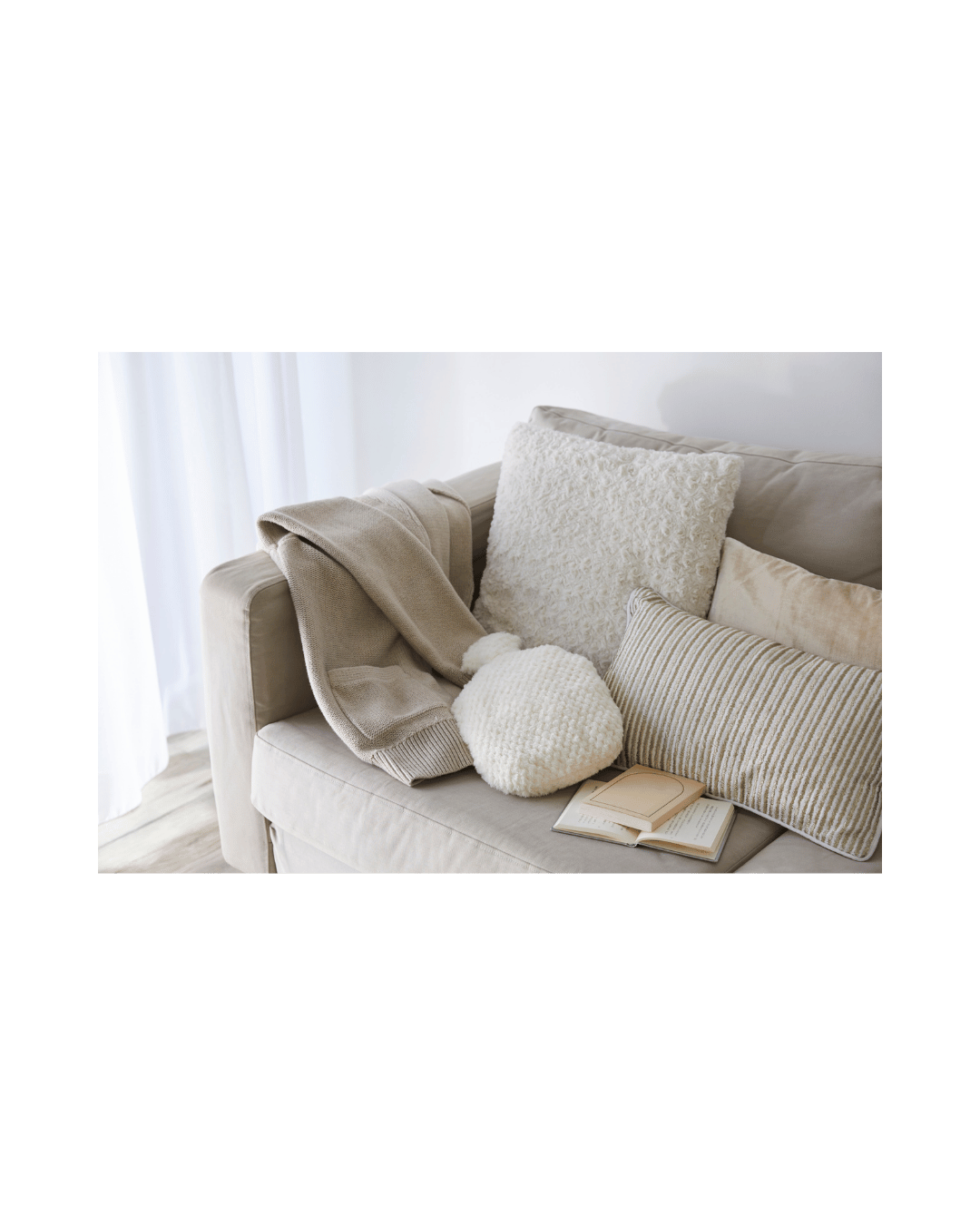 Cream-colored sofa with beige and striped pillows, a beige blanket, a white textured cushion, a fluffy white round cushion, and an open book, in a bright room with white curtains.