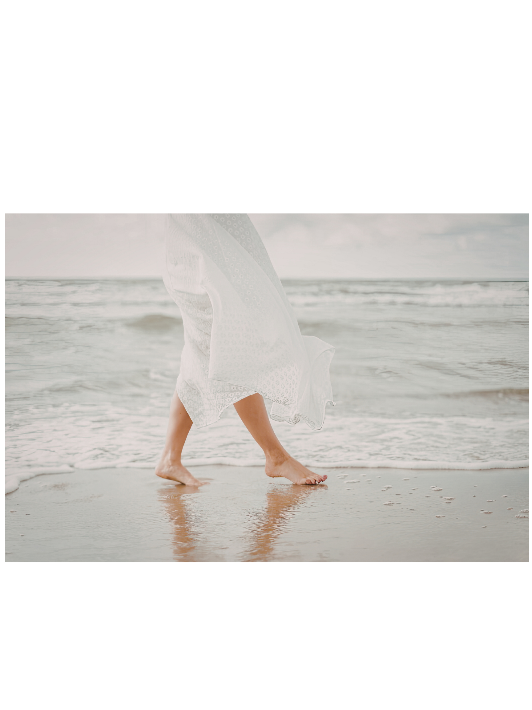 Person walking barefoot along the shoreline, holding a patterned umbrella, with waves and cloudy sky in the background.