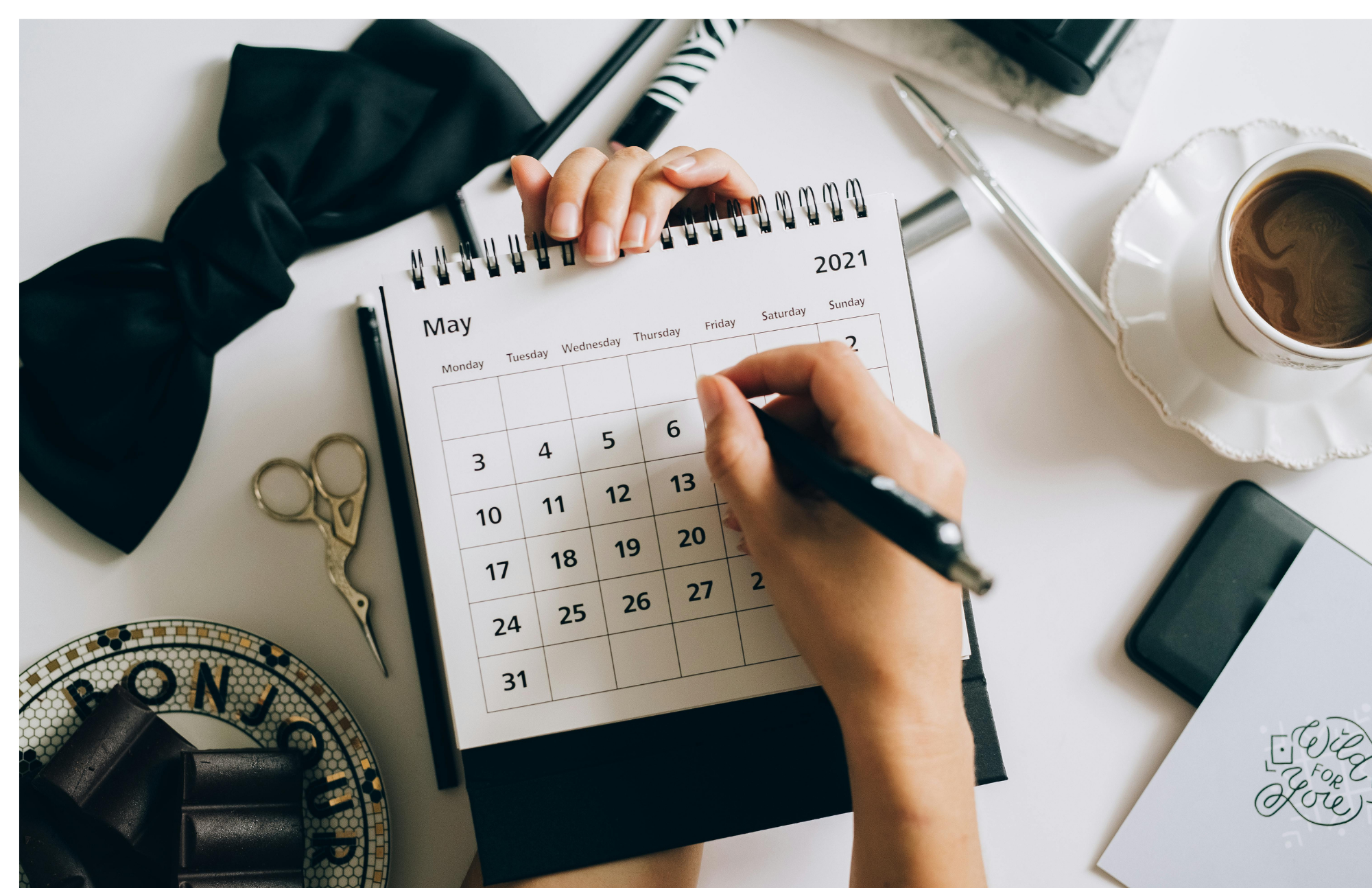 A person is writing in a May 2021 calendar on a desk. Surrounding items include a cup of coffee, scissors, a pen, a notebook, and decorative items.