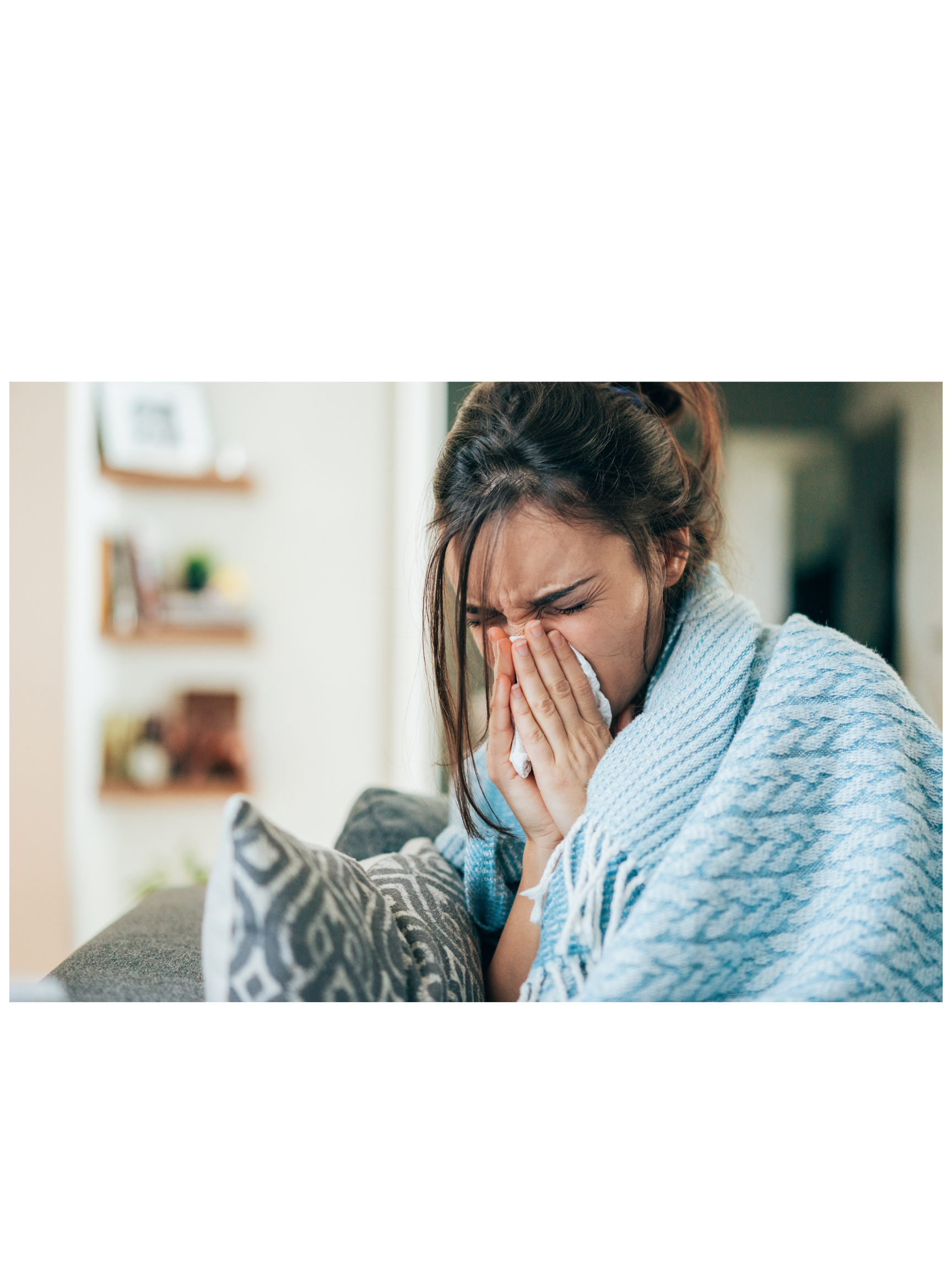 A woman sitting on a couch, holding a tissue to her face, appears upset or crying. She has dark hair and is wearing a light-colored, striped sweater. The background shows a blurred living room with shelves and picture frames.
