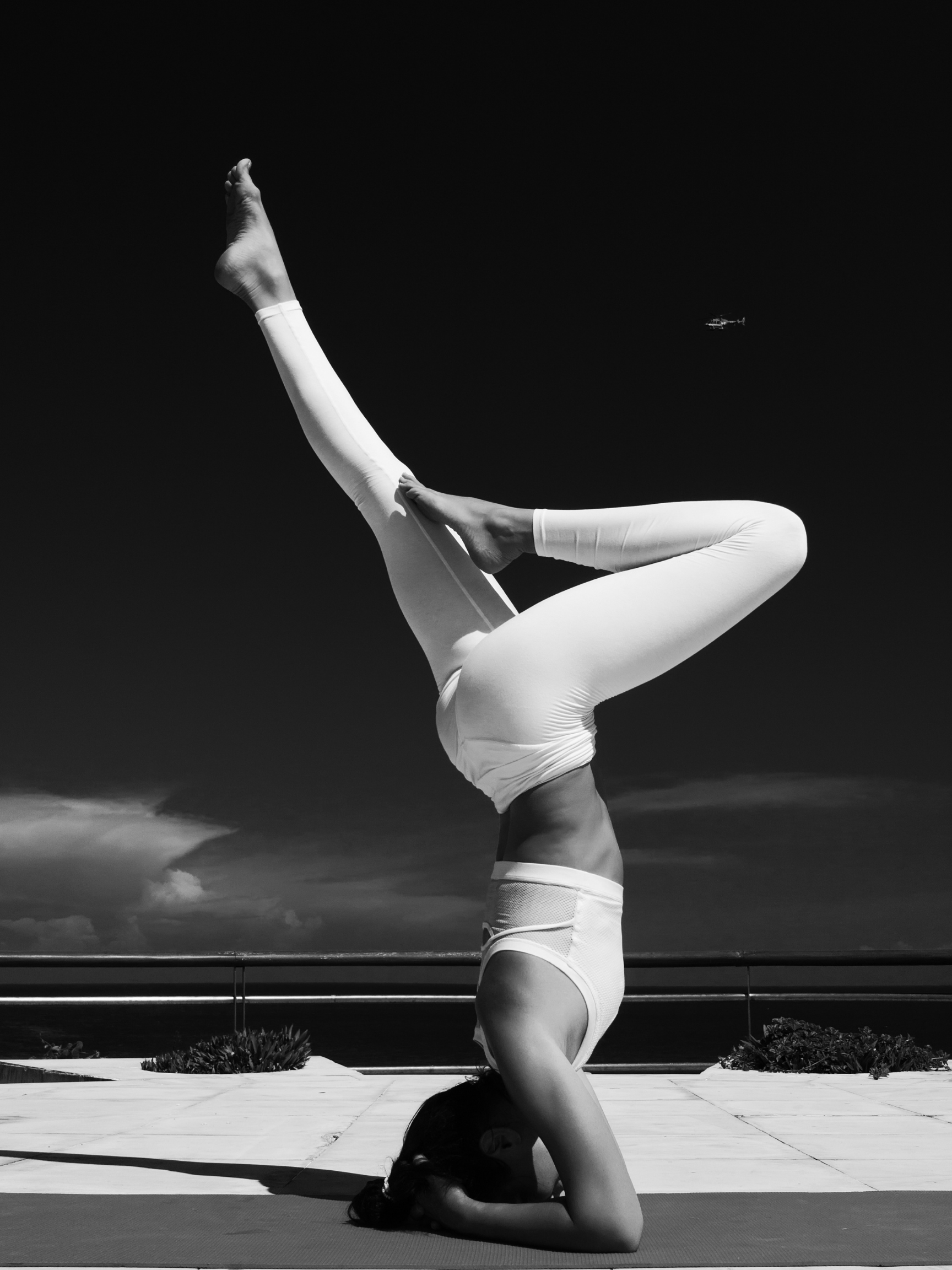 A person performing a headstand yoga pose outdoors, wearing white leggings and a sports bra, with the background of a clear sky and a distant helicopter.