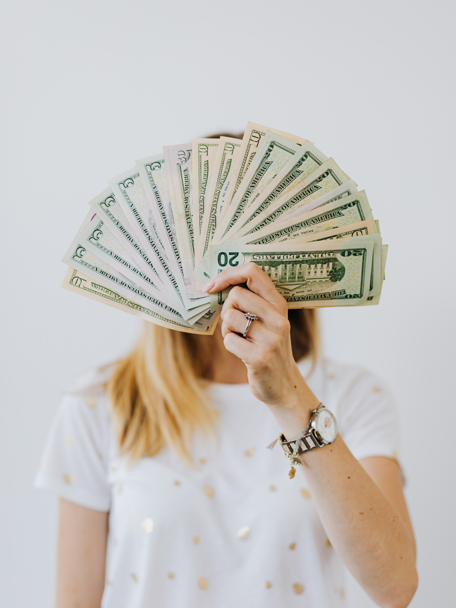 Person holding a fan of US dollar bills in front of their face, wearing a watch and bracelet, with a white and gold polka dot shirt and blurred background.