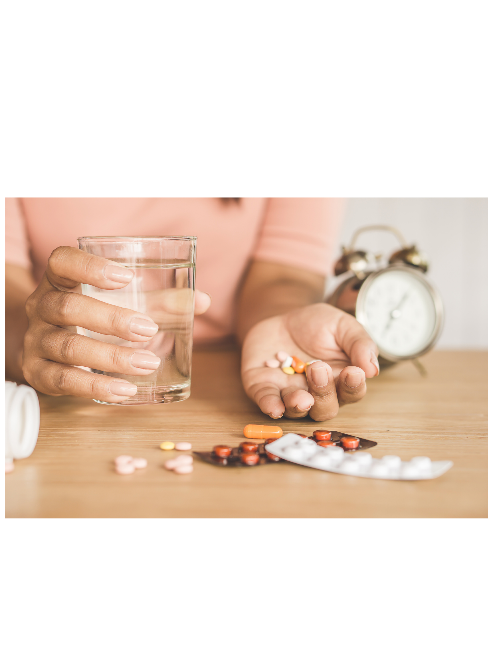 Close-up of a person holding a glass of water in one hand and a few pills in the other, with a vintage alarm clock in the background and pills scattered on a wooden surface.
