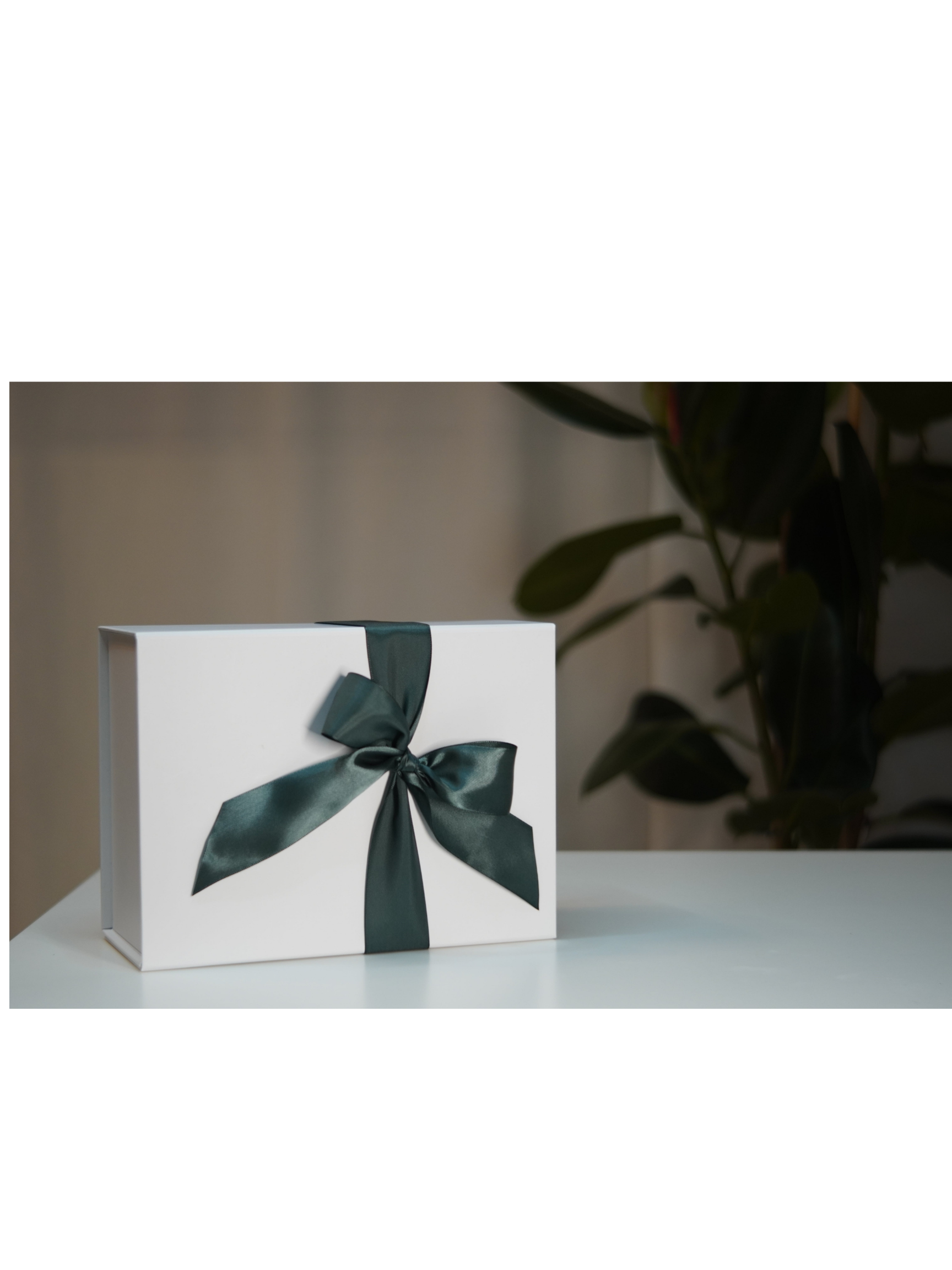 A white gift box with a dark green satin ribbon tied in a bow, sitting on a white table in front of a background of green leafy plants.