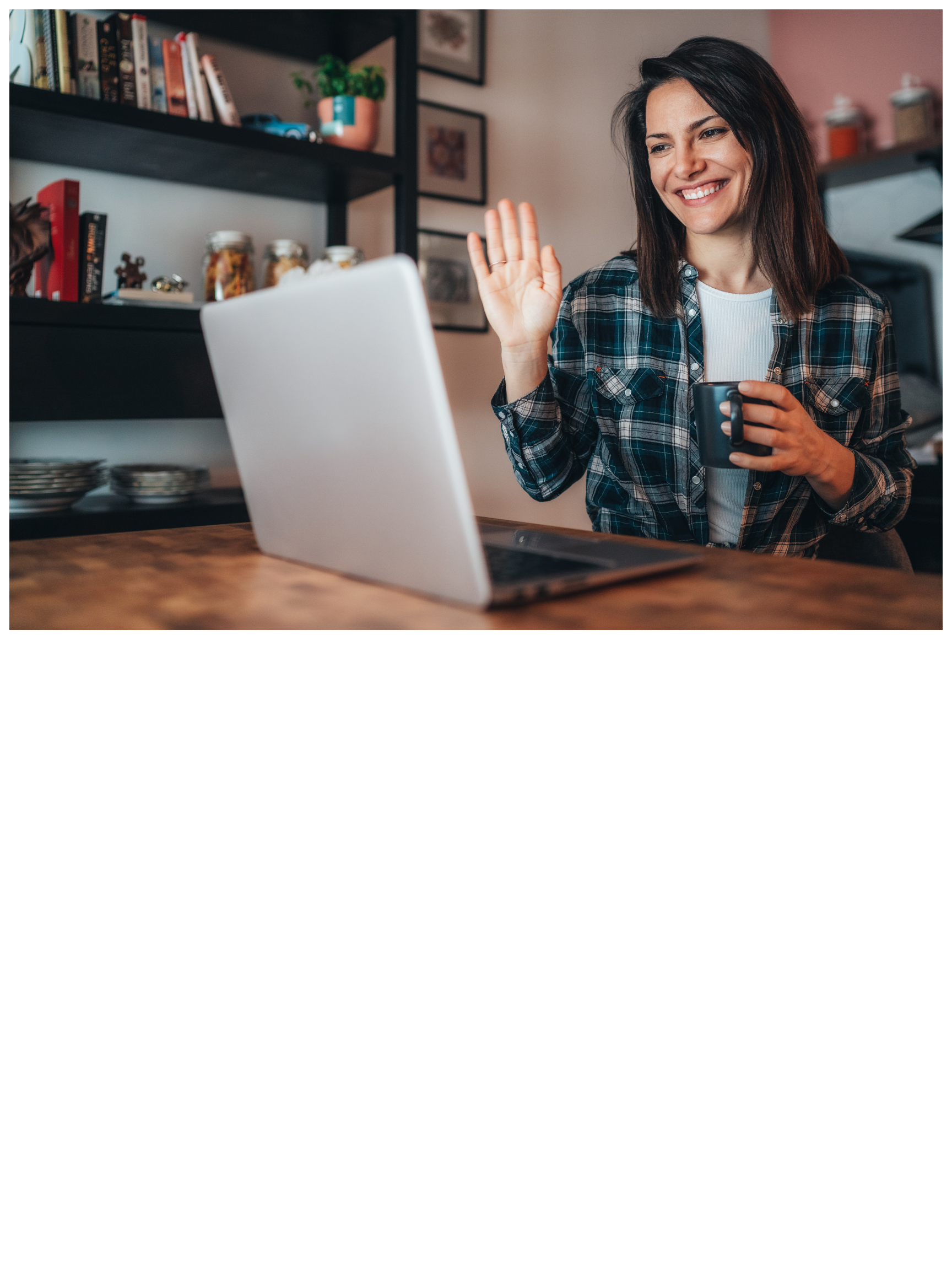A woman with shoulder-length dark hair smiling and waving while holding a black mug. She is sitting at a wooden table with a silver laptop in front of her. The background features black shelves with books, decorative jars, and framed pictures, creating a cozy, home office or kitchen setting.