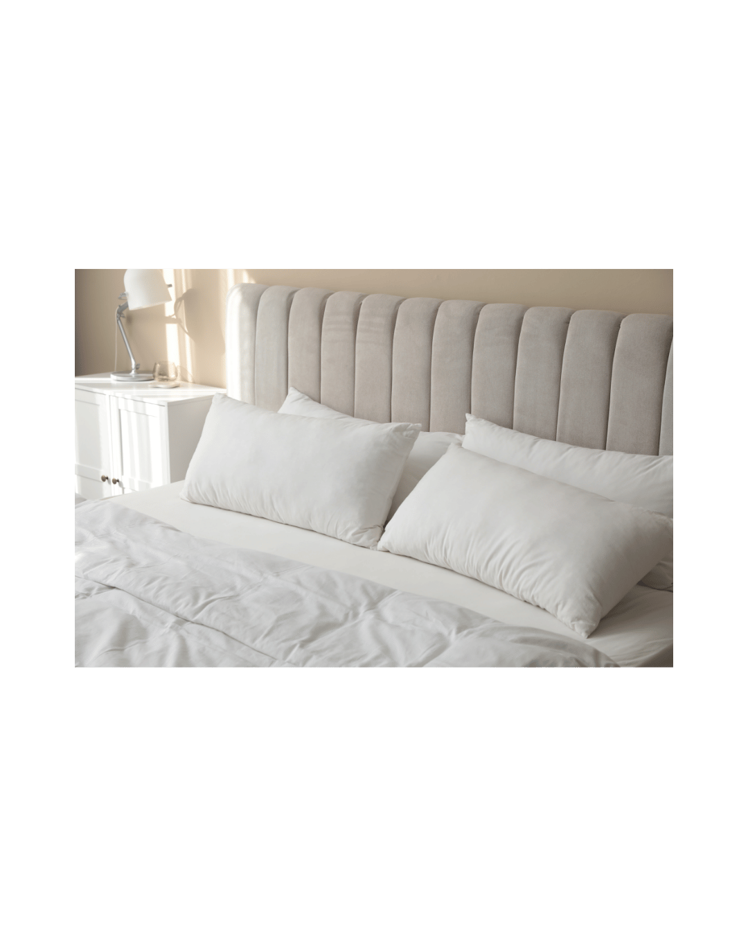 A neatly made bed with an upholstered cream-colored headboard, white pillows, and a white duvet in a bright, well-lit bedroom.