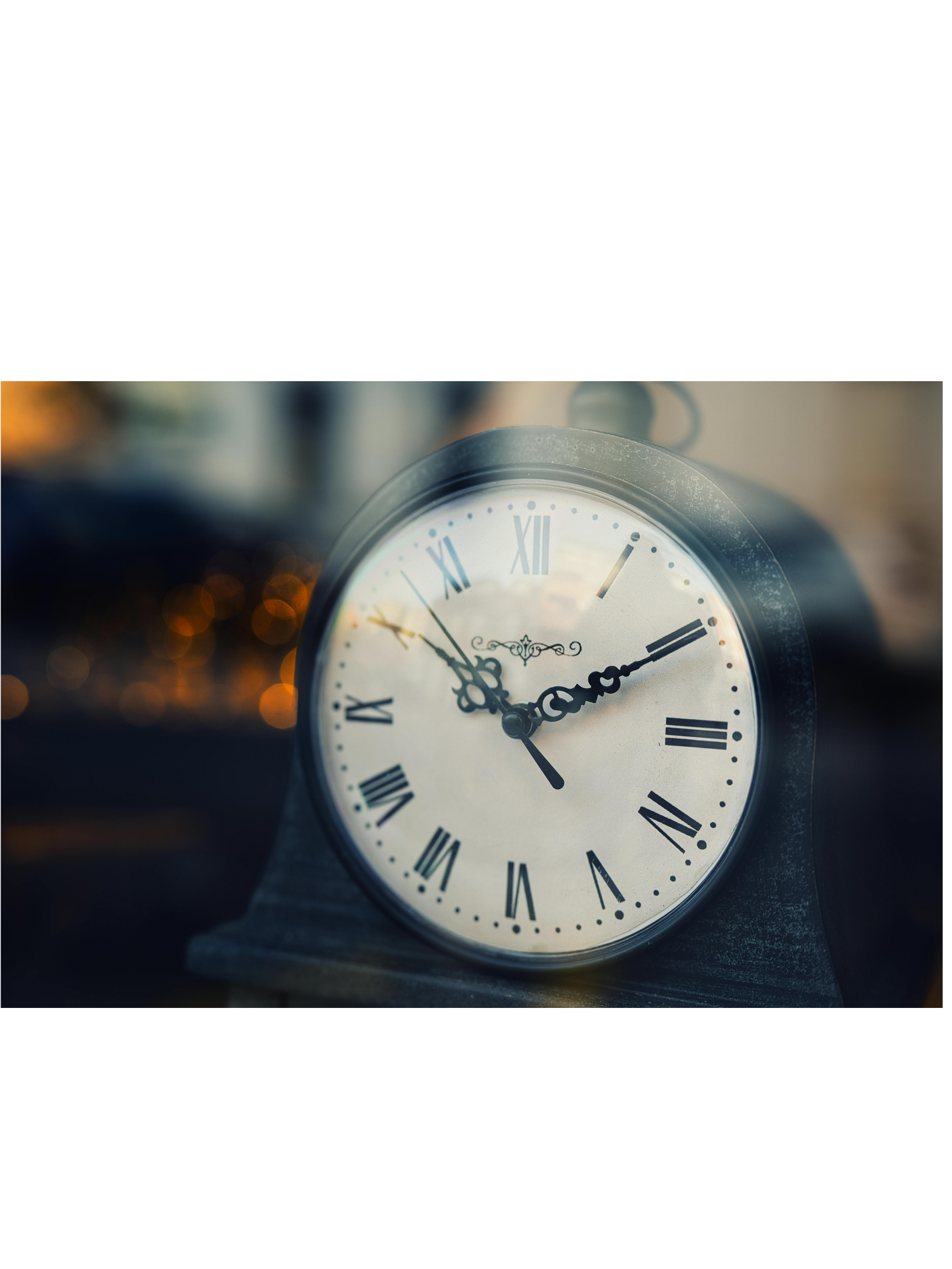 Close-up photograph of an analog clock with Roman numerals showing the time as 10:10. The clock has ornate black hands and a metal frame, with a blurred background featuring warm bokeh lights.