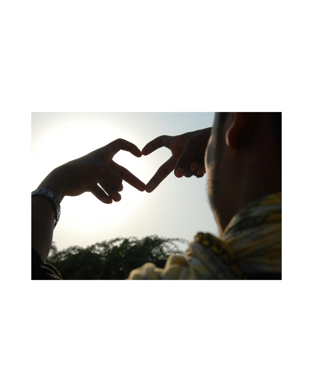 Silhouette of two people forming a heart shape with their hands against the sun, with trees in the background.