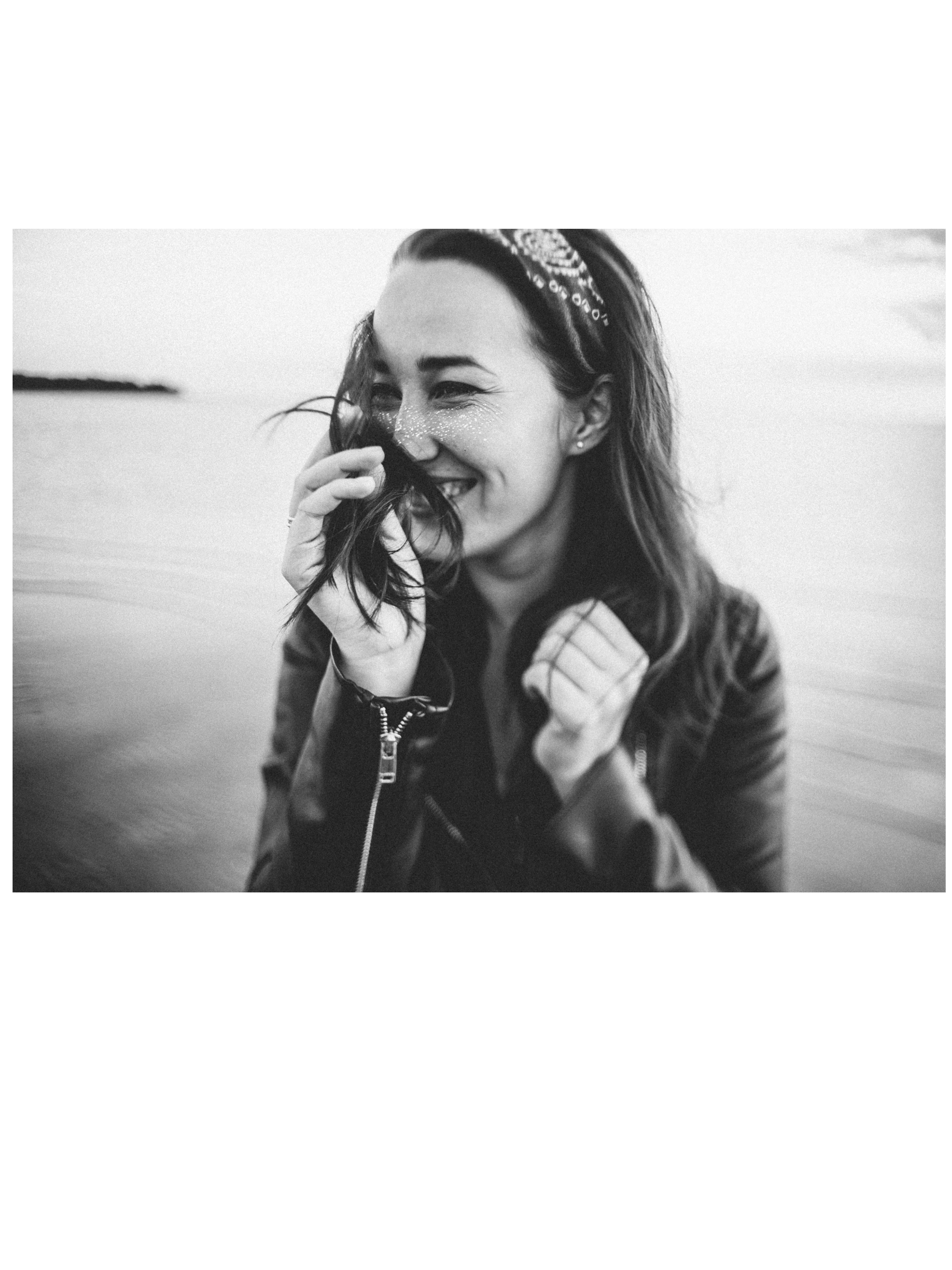 Black and white photo of a woman smiling, with glitter on her nose, wearing a headband, and standing near water.