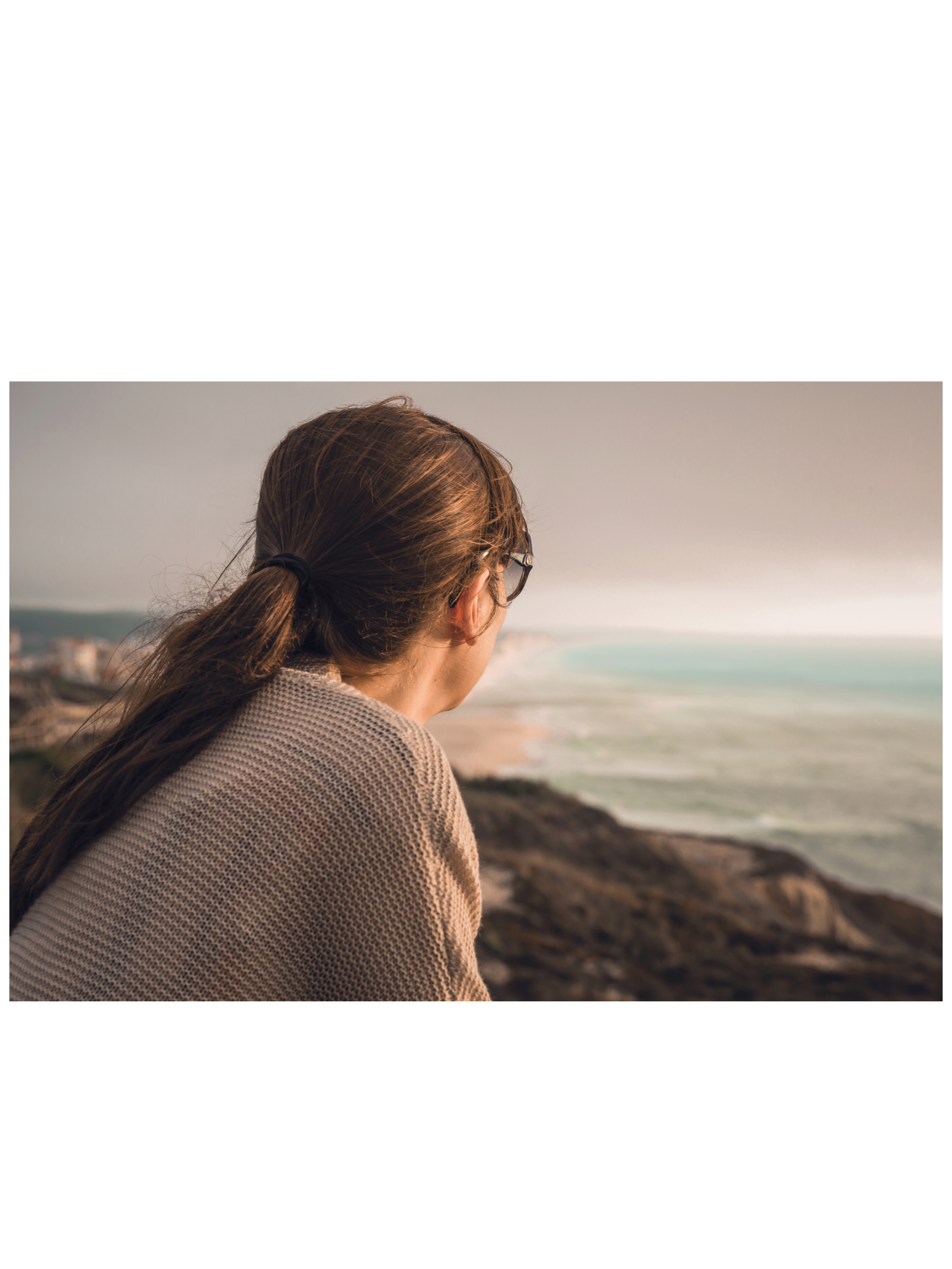 A woman with brown hair tied back, wearing glasses and a beige sweater, sitting on a rock and looking out at the ocean during sunset, with a coastal town in the background.