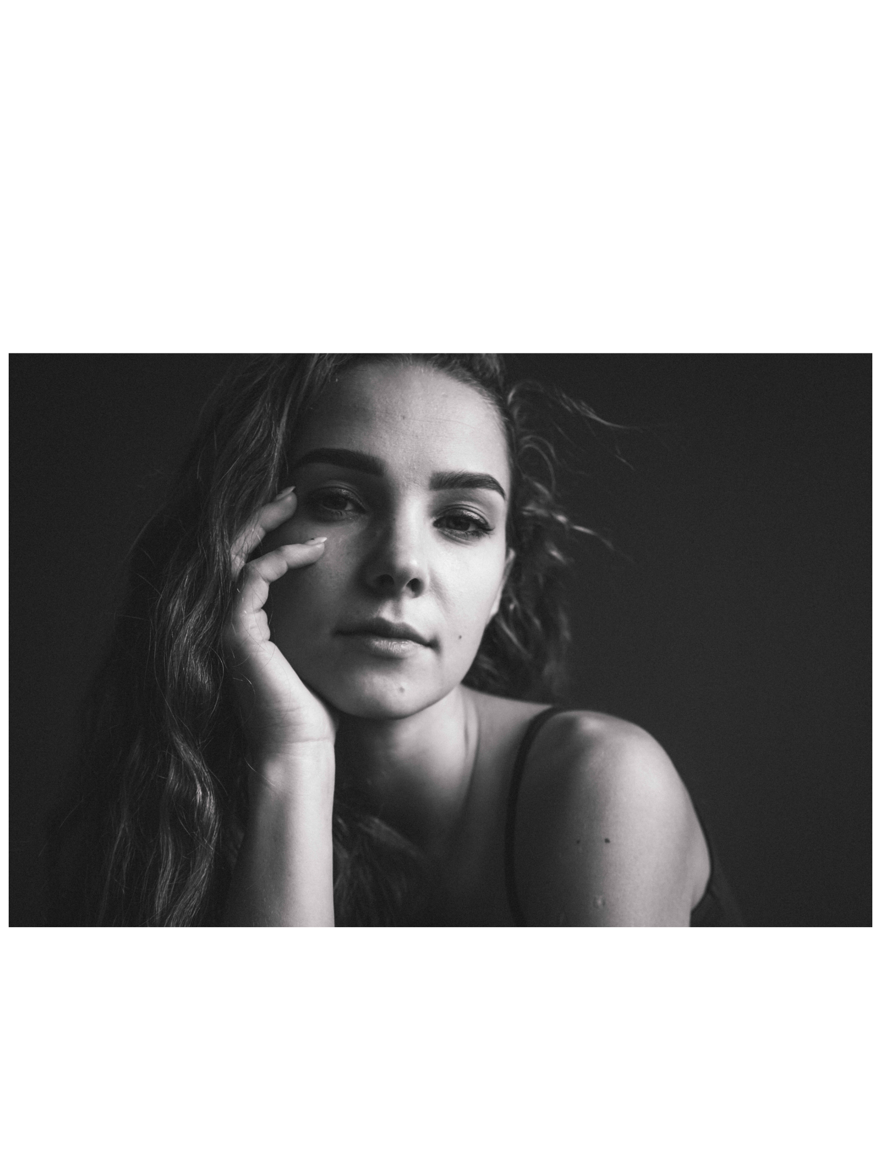 Black and white portrait of a young woman with wavy hair, resting her hand on her face, looking directly at the camera with a calm expression.