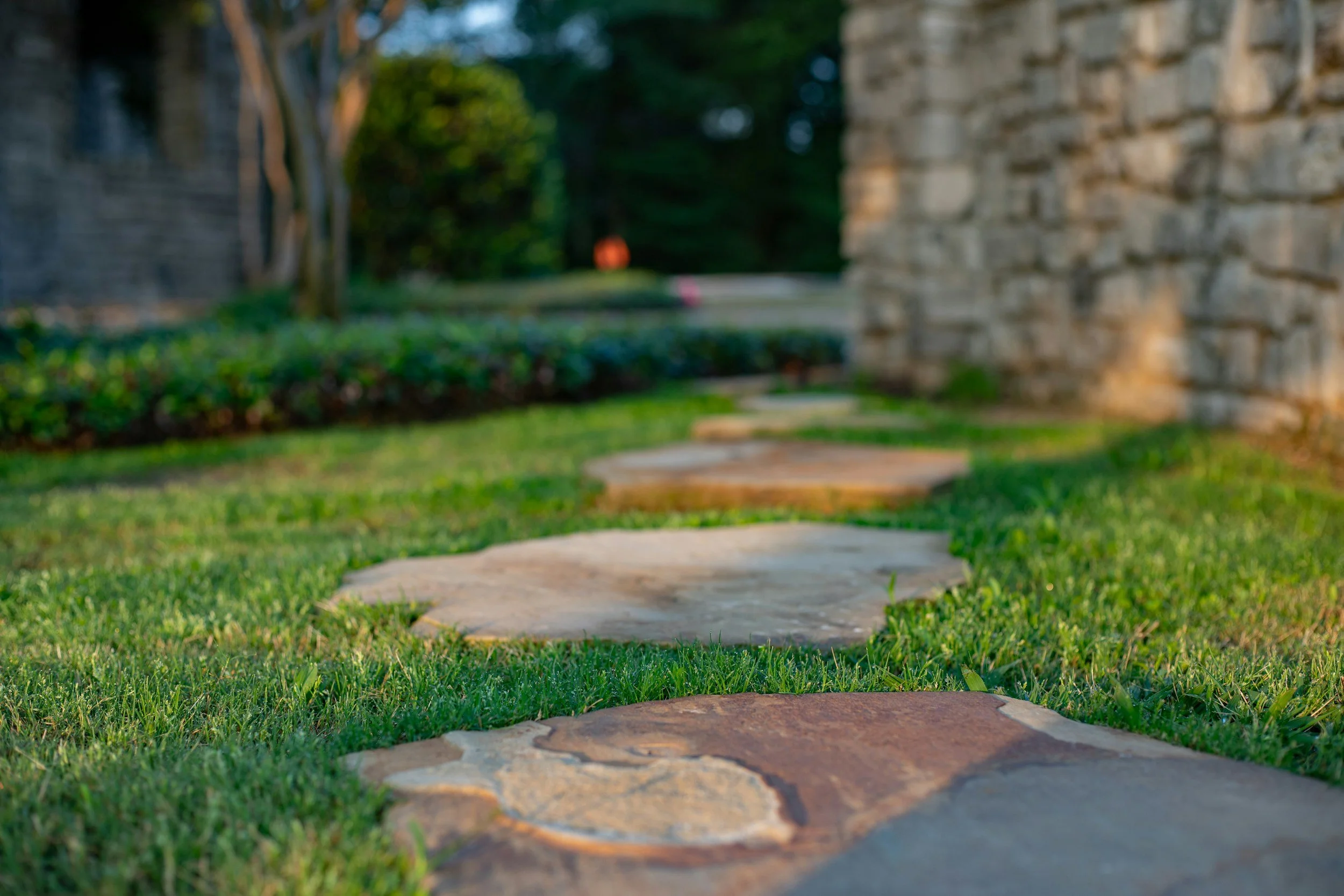 A stone pathway surrounded by green grass, leading toward a blurred background with trees and a brick wall on one side.