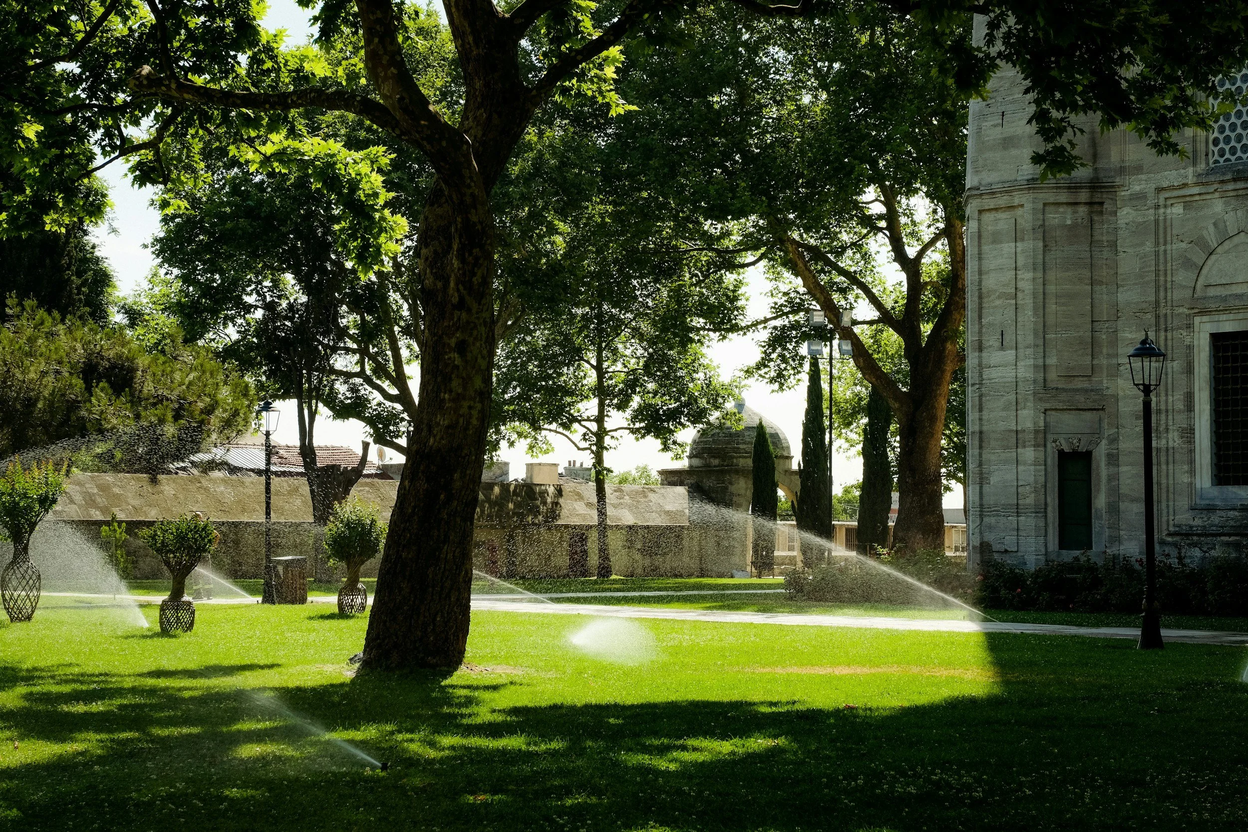 A lush green park with large trees and a stone building, with water sprinklers watering the grass during the daytime.