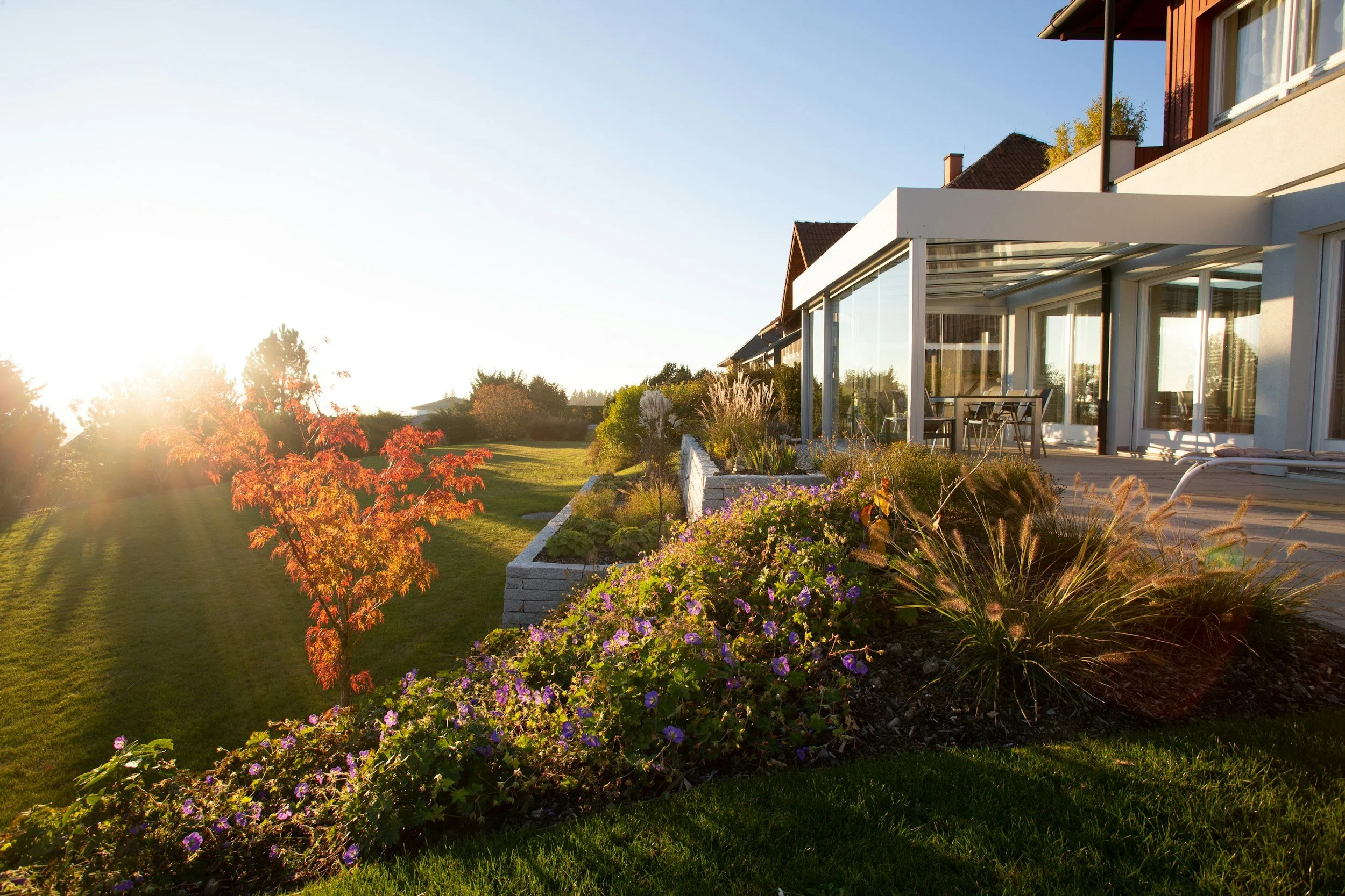 A modern house with large windows and a patio, surrounded by a well-maintained lawn and colorful flower garden, illuminated by sunlight.