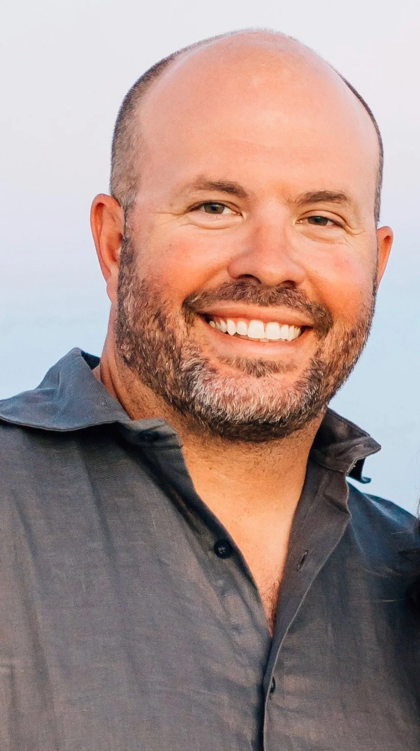 Smiling man with a shaved head and beard wearing a dark collared shirt, outdoors with a blurred sky background.