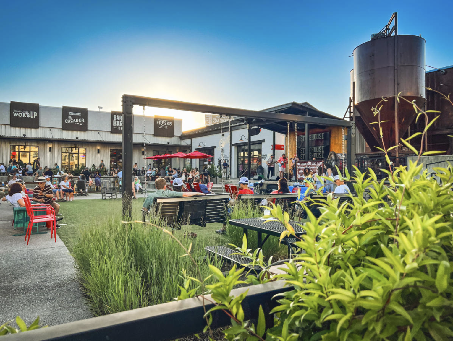 Outdoor concert at a restaurant with audience seated on chairs and benches, live band playing on stage, tables with red umbrellas, modern industrial-style building in the background, and a large metal silo on the right. The scene is during sunset wit