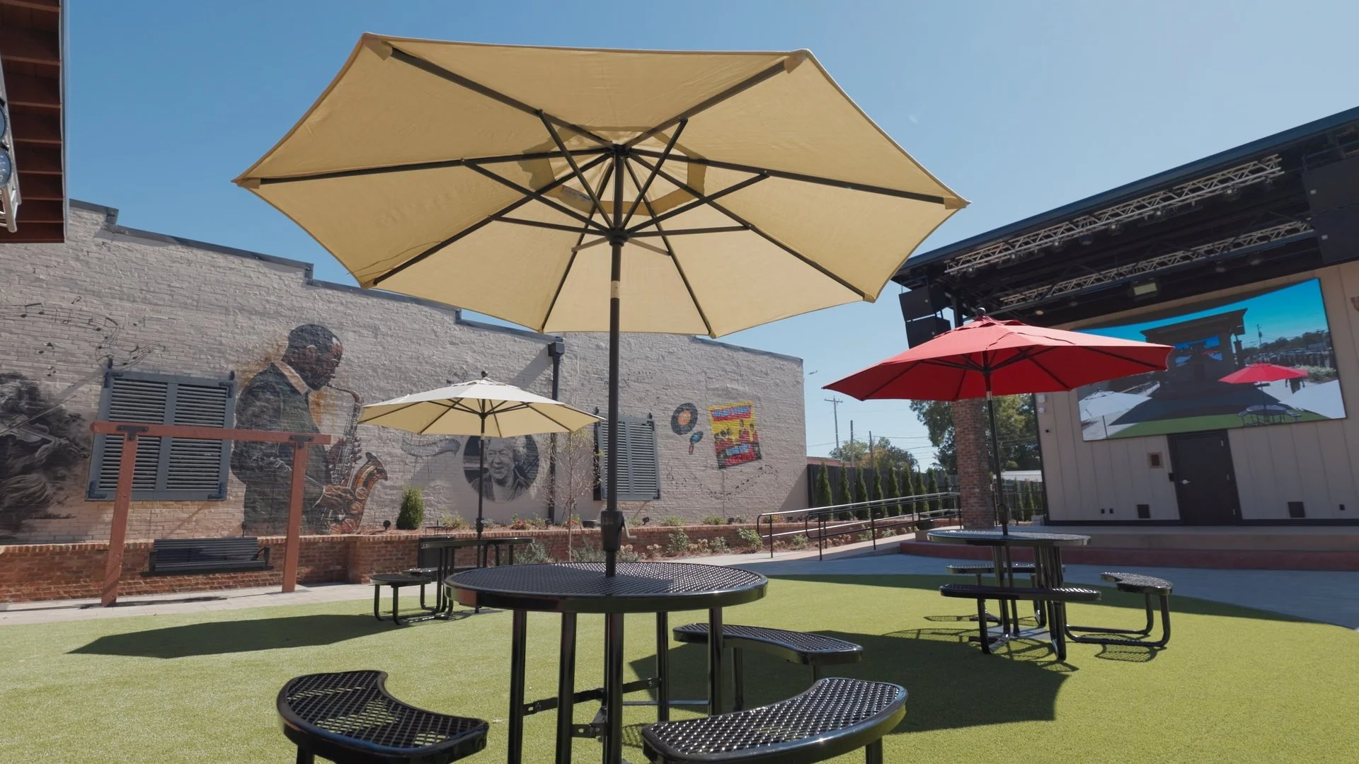 Outdoor area with round black metal tables and benches, large yellow and red patio umbrellas, a brick wall with a mural of a man playing saxophone and people smiling, a stage with a large screen, and a grassy ground under a clear blue sky.