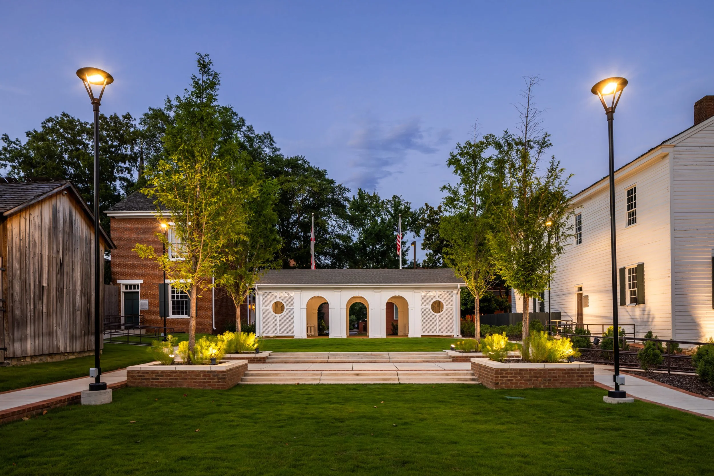 Nighttime view of a well-lit garden with trees, a white gazebo, and surrounding buildings, illuminated by streetlights and landscape lighting.