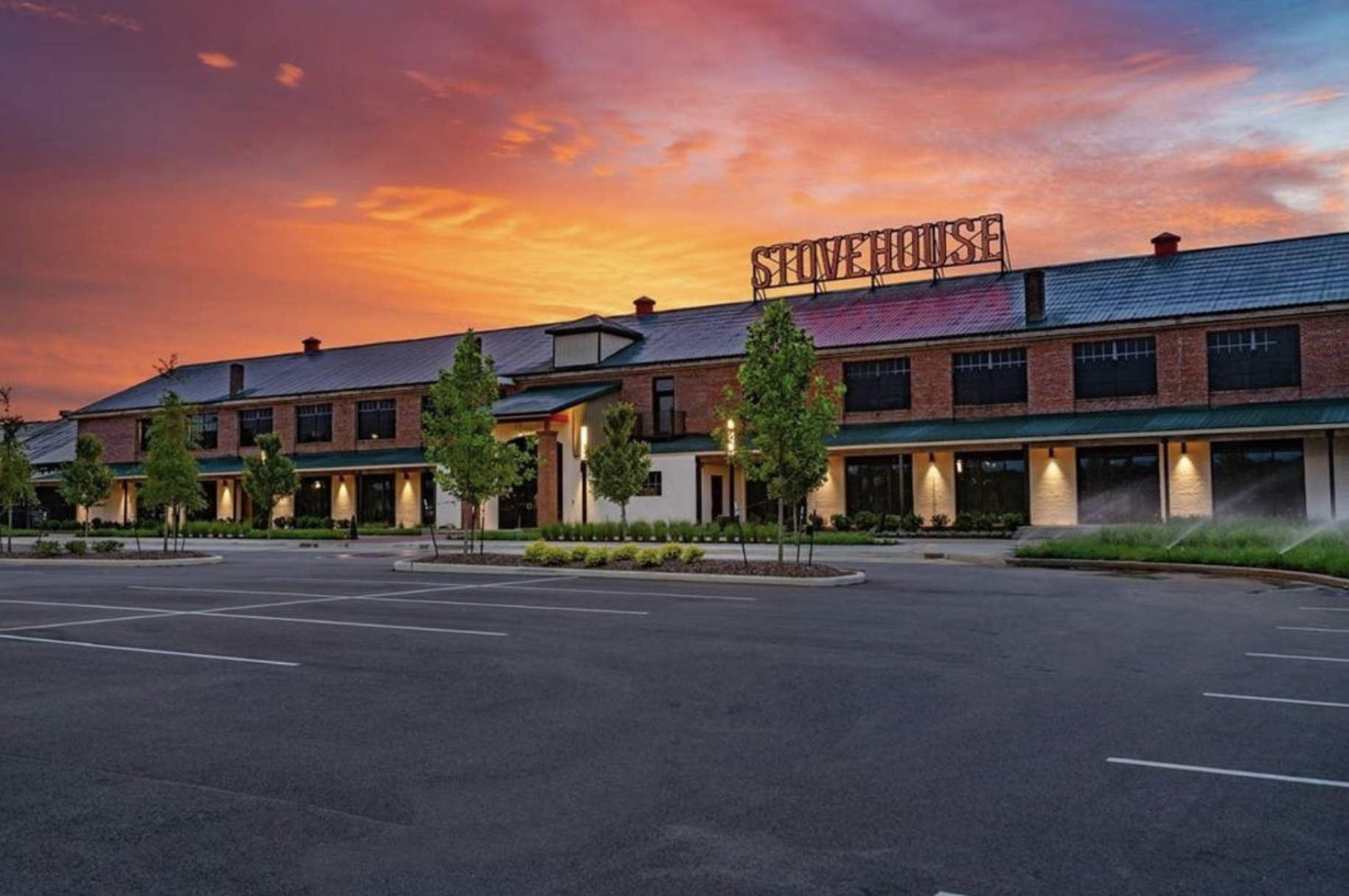An empty parking lot in front of a modern restaurant or commercial building with a sign that says "Stovehouse". The building has a brick facade, large windows, and outdoor lighting. The sky is colorful with a sunset or sunrise, featuring orange, pink, and purple hues. There are small trees and landscaped areas around the building.