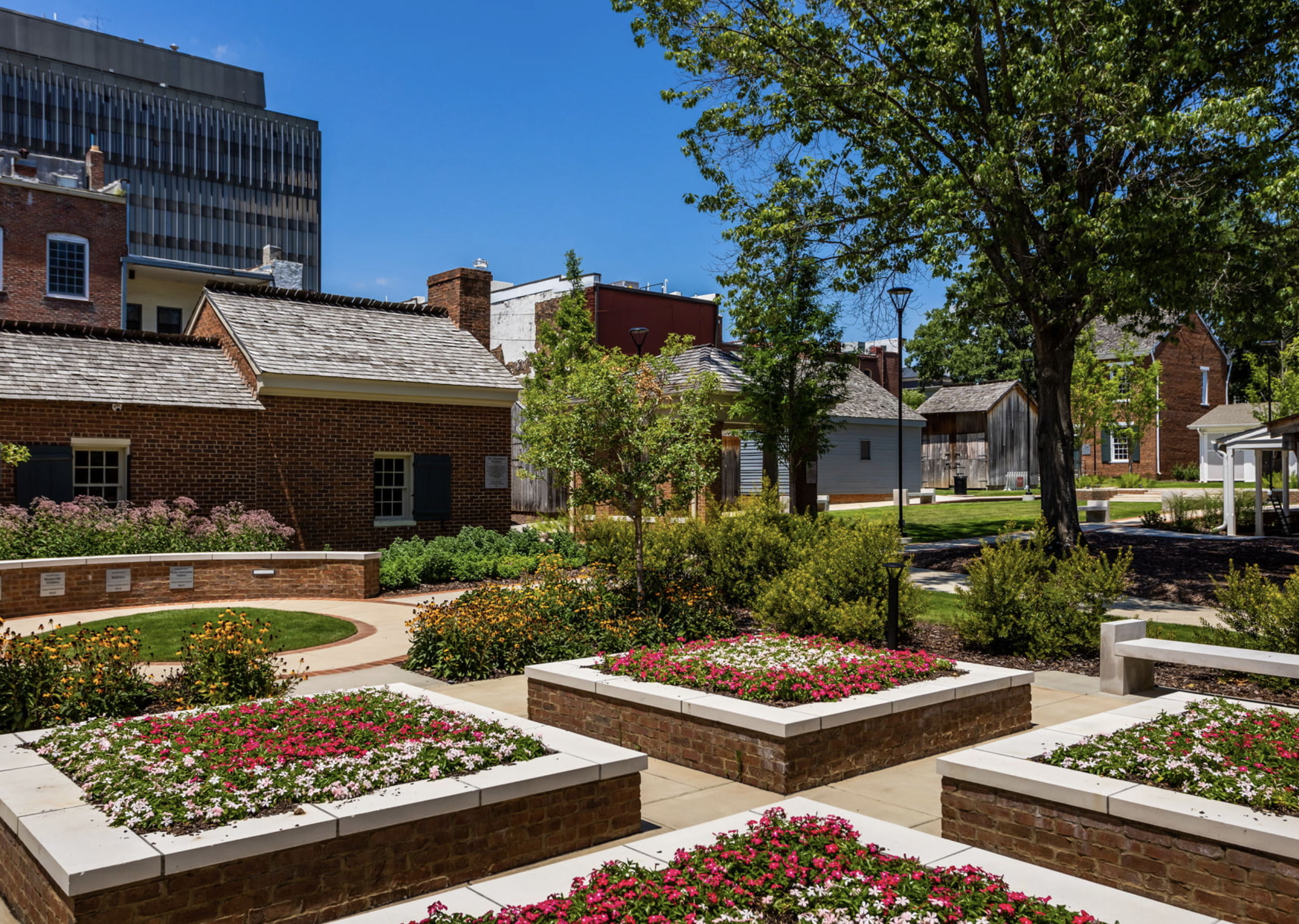 A landscaped urban park with flower beds, brick walls, trees, and city buildings in the background on a sunny day.