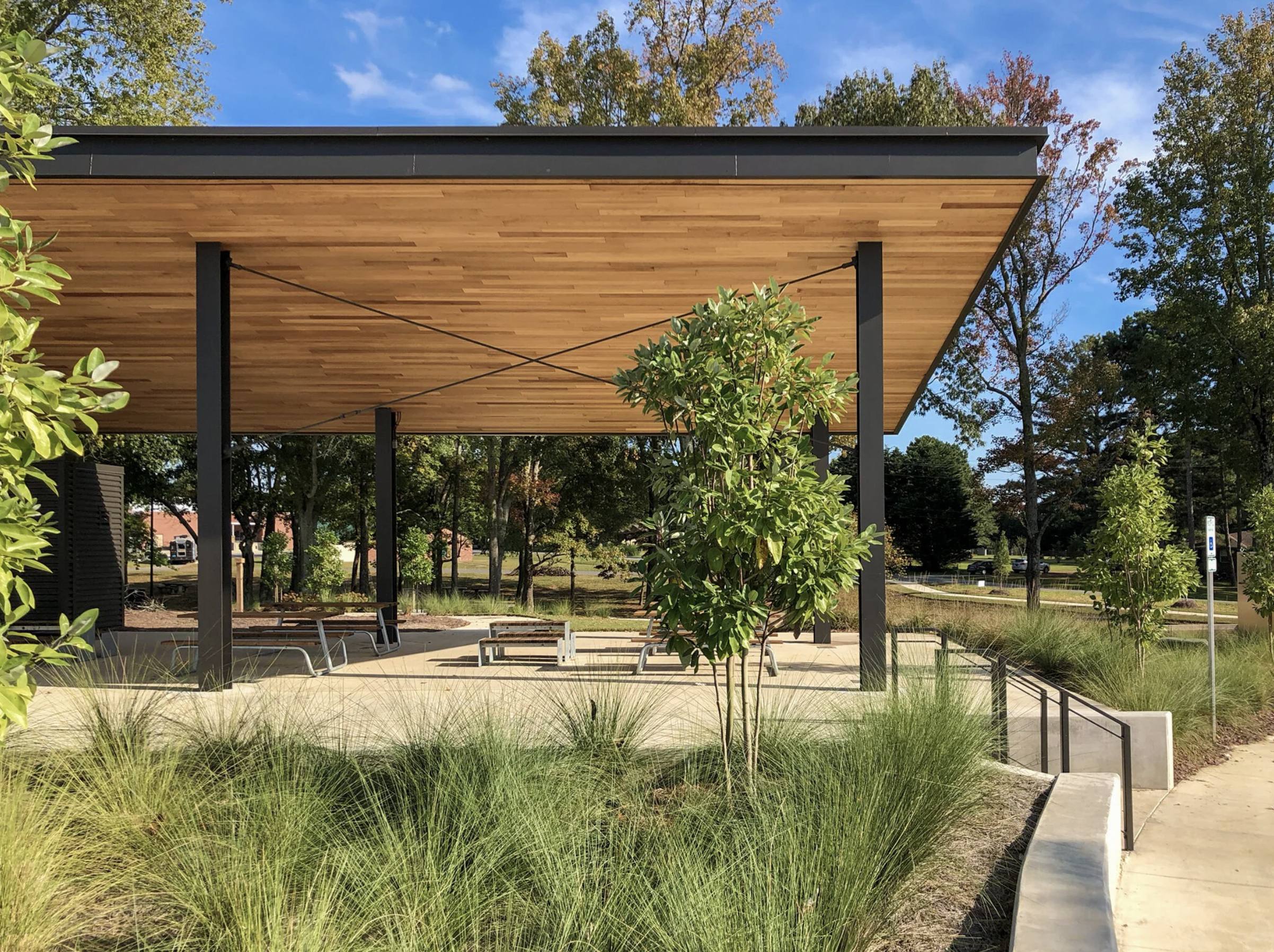 A modern outdoor shelter with a wooden ceiling, black support columns, surrounded by trees, shrubs, and a paved area with benches.