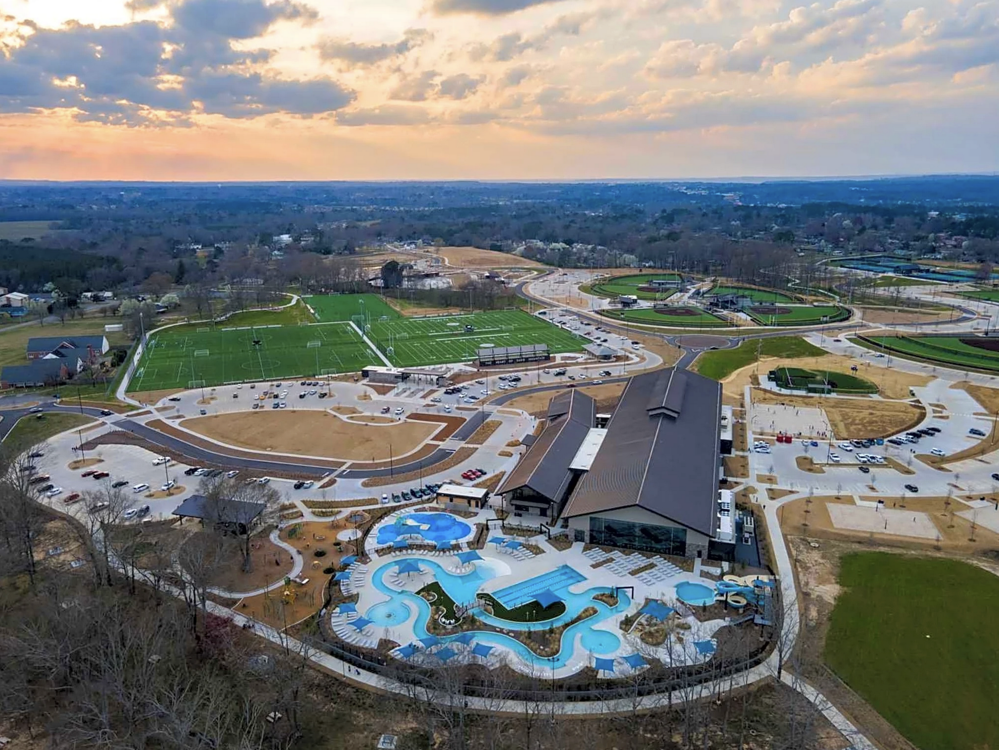 Aerial view of a sports complex and water park with multiple pools, lazy rivers, and colorful umbrellas, surrounded by parking lots and a hilly landscape with trees.