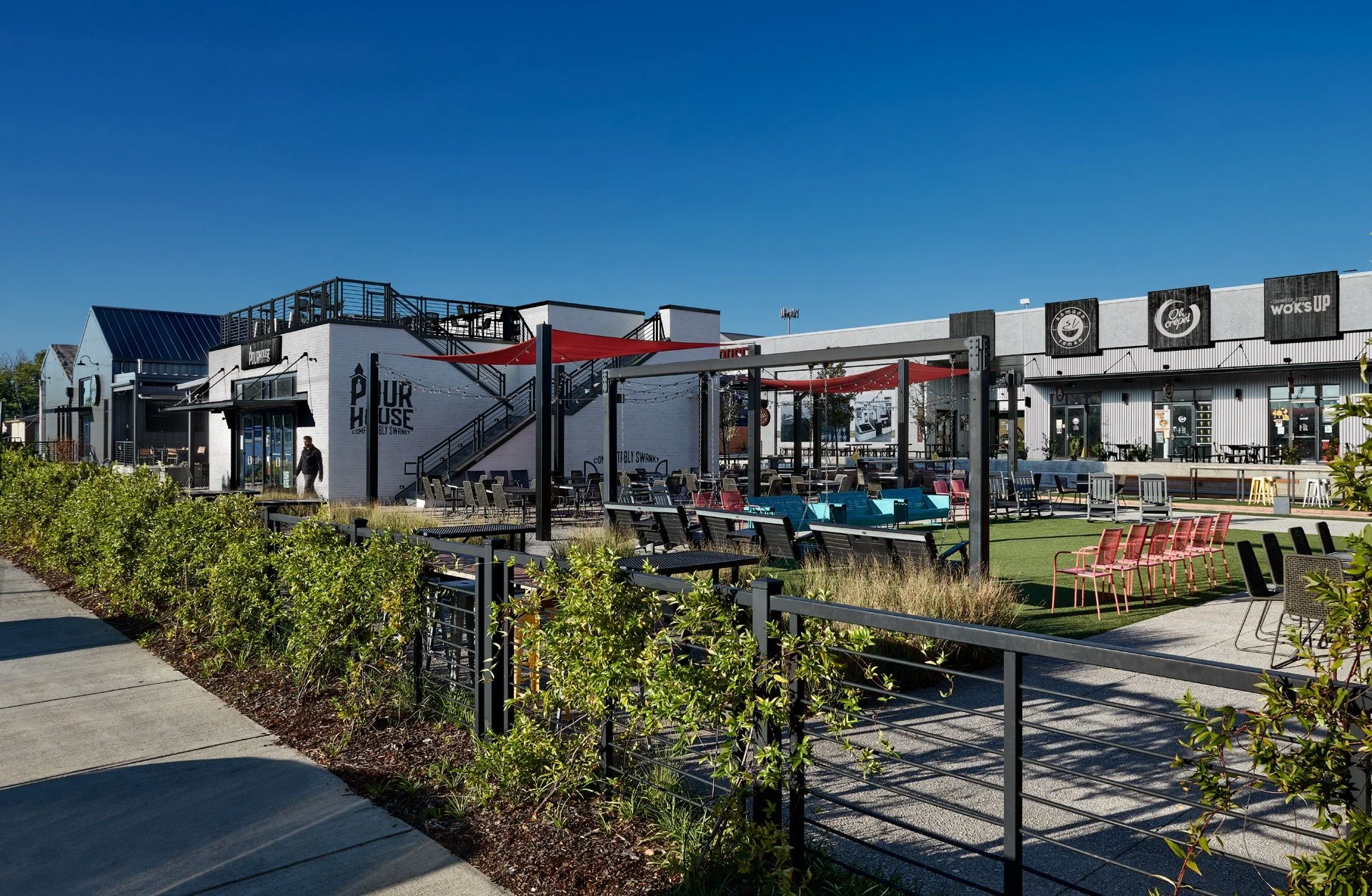 Outdoor seating area with colorful chairs and tables, greenery, and a white building with black signage under a clear blue sky.