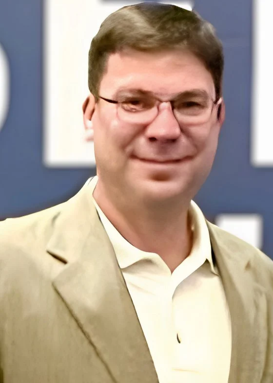 A man with short brown hair, glasses, and a light beard, wearing a beige blazer and a white shirt, smiling at the camera in front of a blue background with white letters.