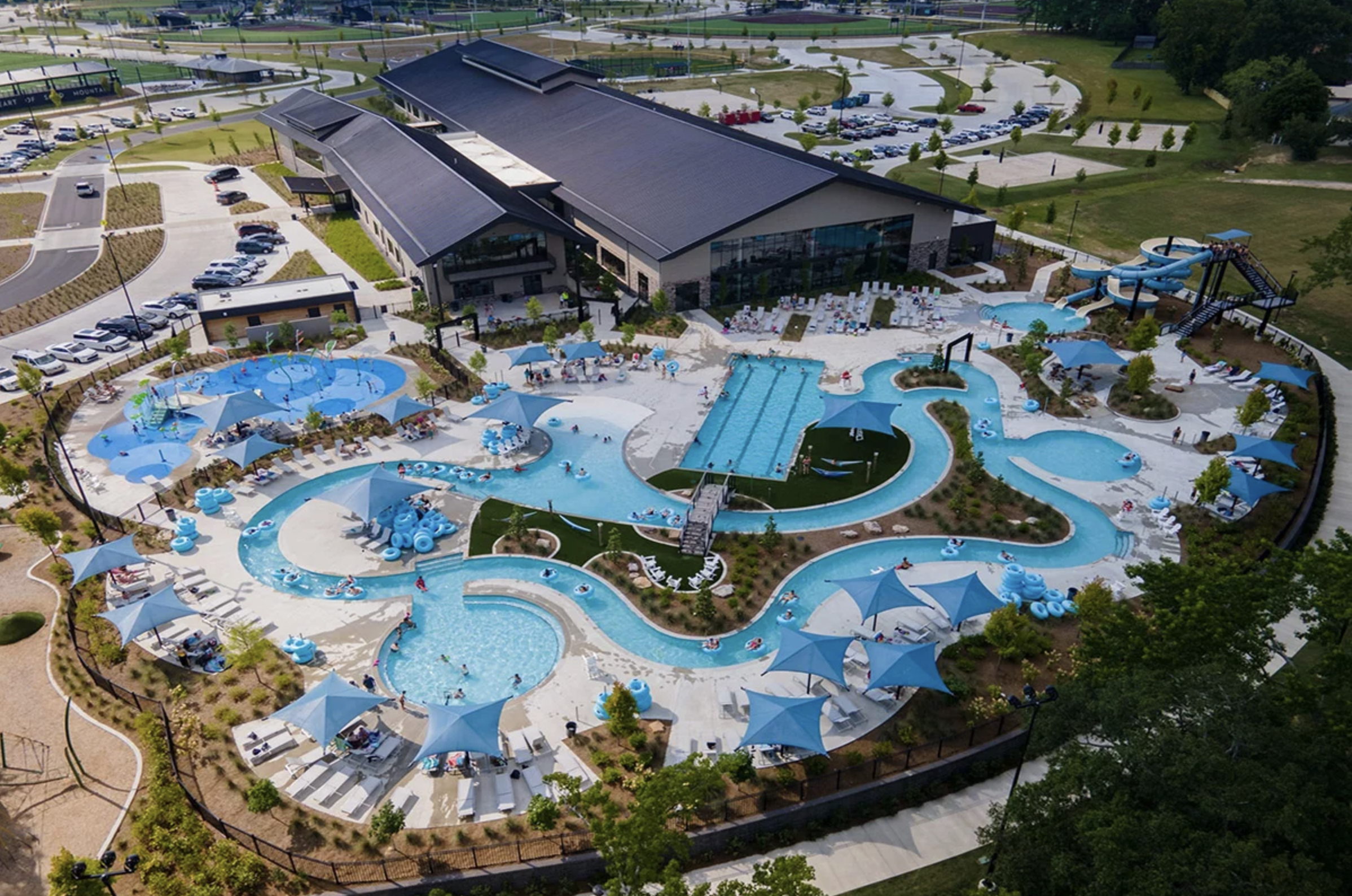 Aerial view of a water park featuring multiple pools, water slides, lazy river, shaded lounging areas with umbrellas, and a large building in the background, surrounded by parking lots and green spaces.