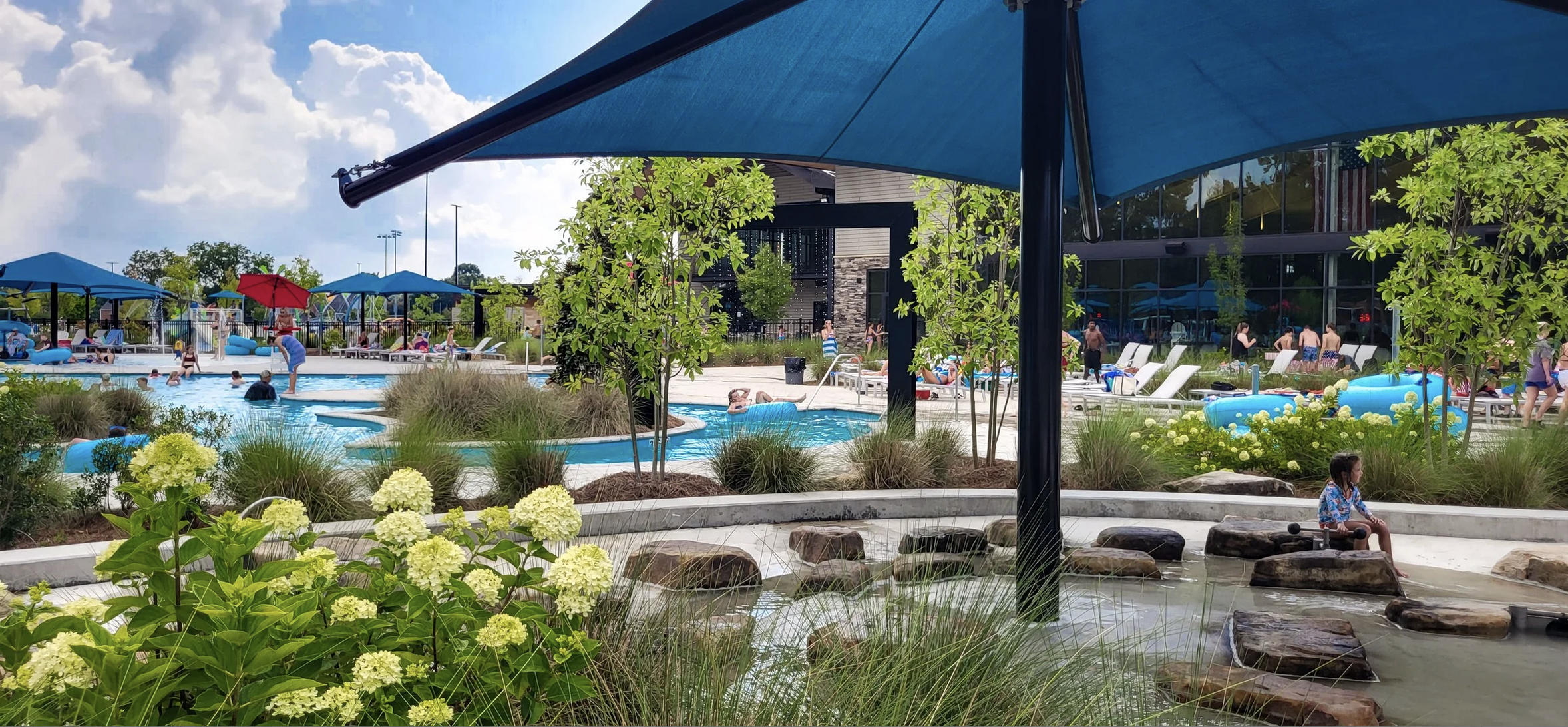 People enjoying a water park with swimming pools, water slides, shaded areas, and landscaping with plants and flowers on a sunny day.