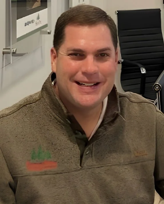 A man with short brown hair smiling at the camera, wearing a brown jacket with a small green and red logo on the left side, sitting in an office or conference room.