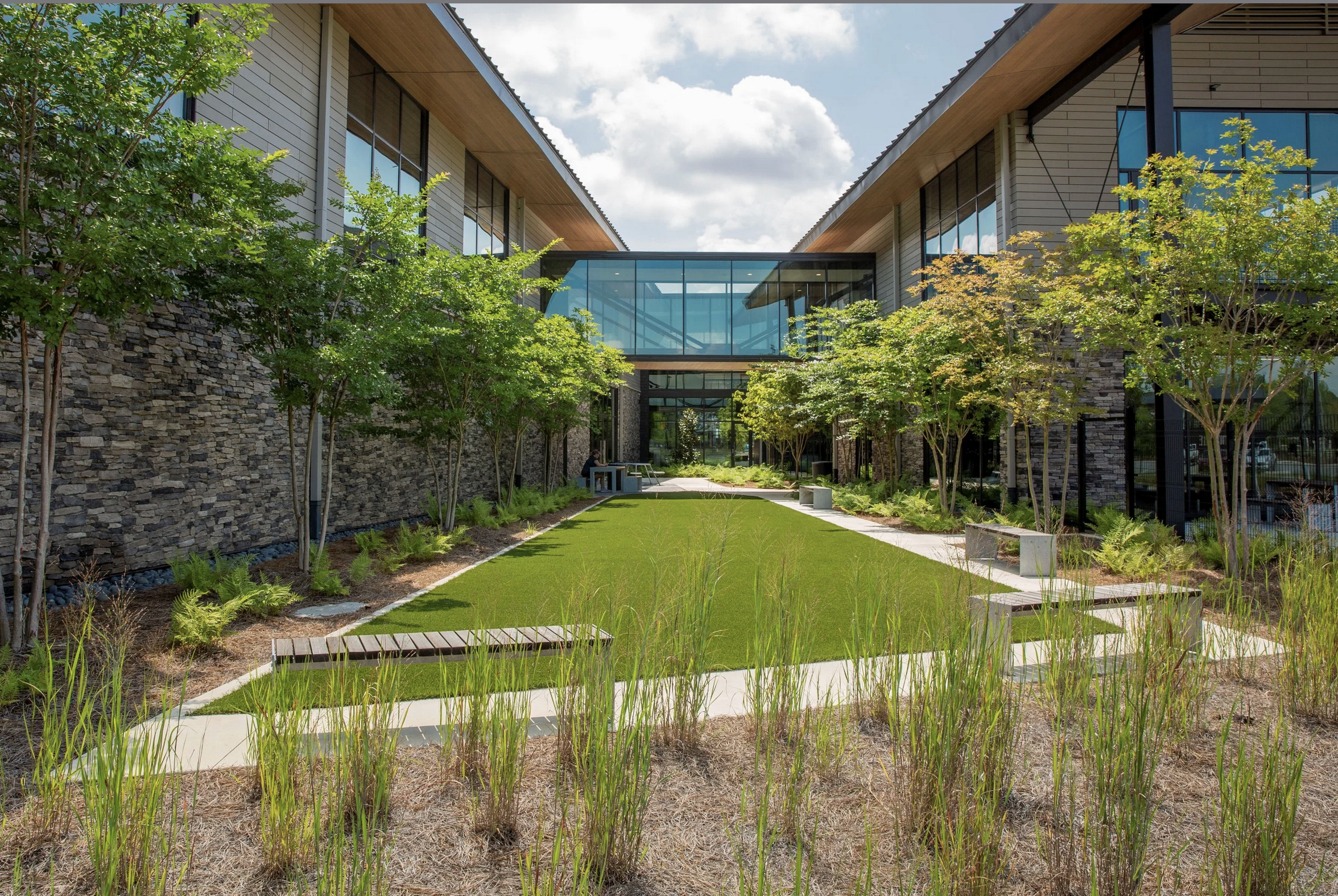 Modern building with glass walkways connecting two structures, and landscaped courtyard with trees, grass, and pathways.