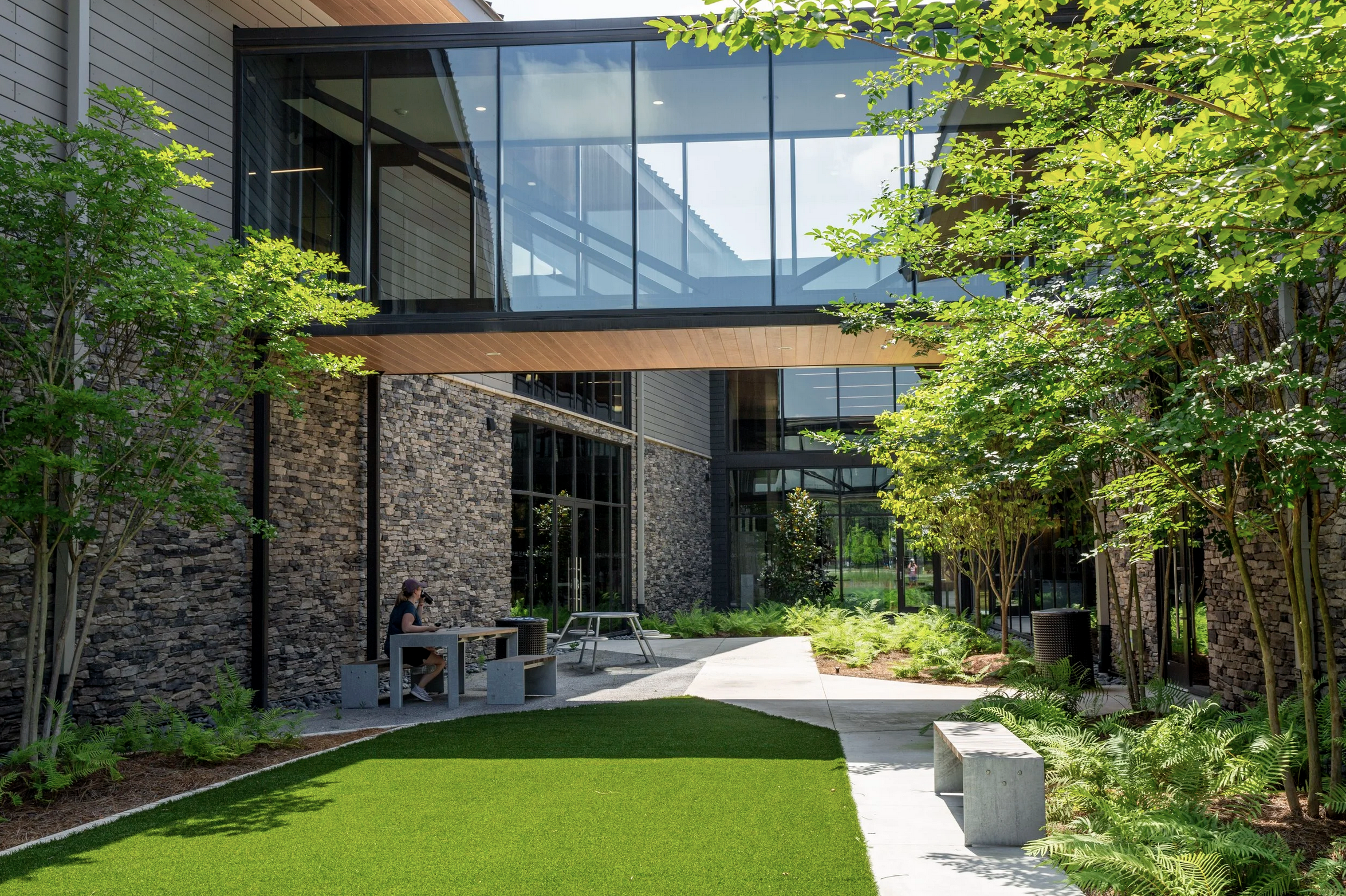 A modern building with large glass windows, stone walls, and a wooden overhang, surrounded by green trees, plants, and a grassy area. There is a person sitting at an outdoor table.