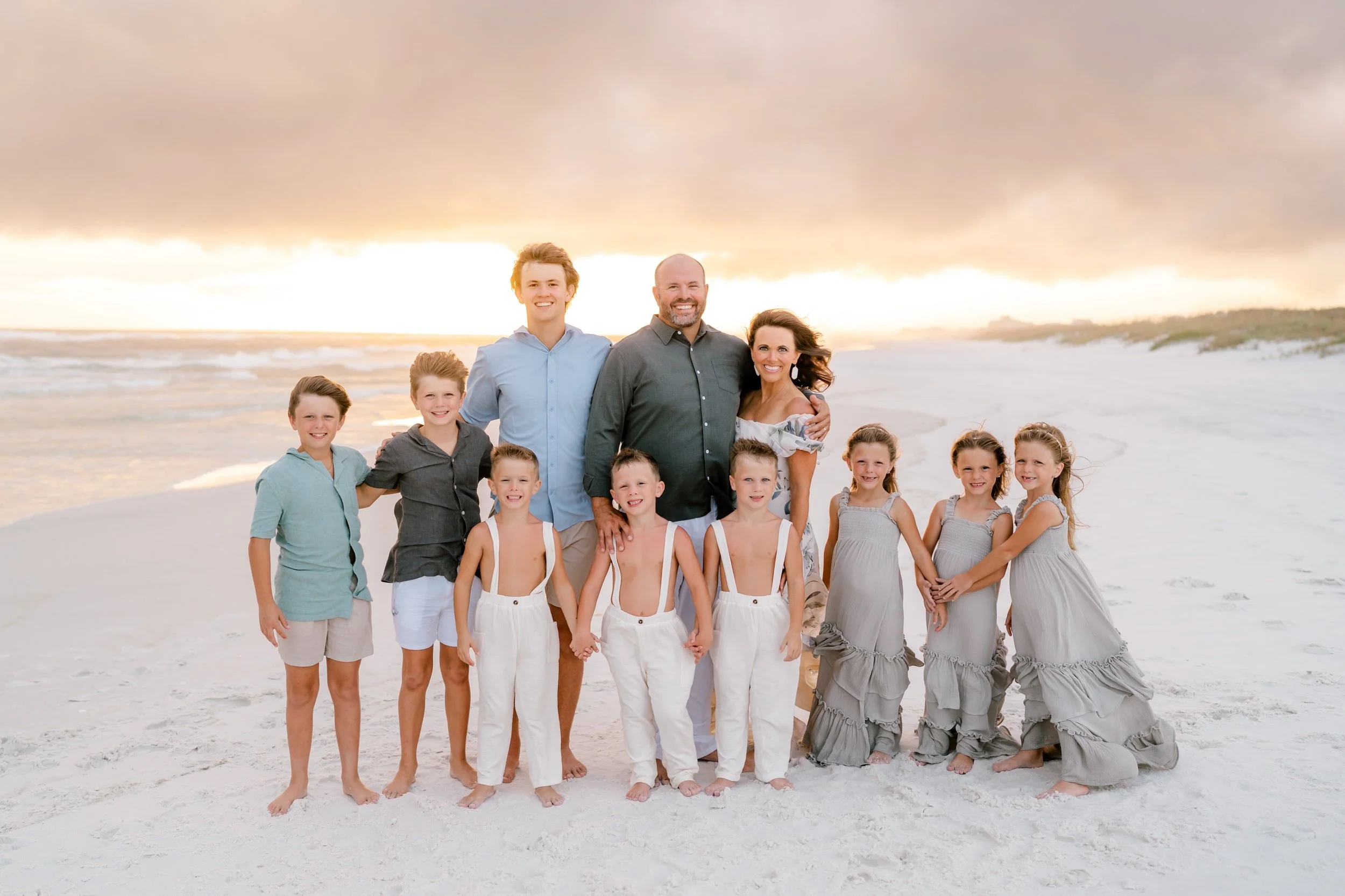 A large family group standing on a sandy beach at sunset, smiling at the camera. The group includes adults and children dressed in light, casual clothing. The ocean is visible in the background under a sky with dramatic clouds.