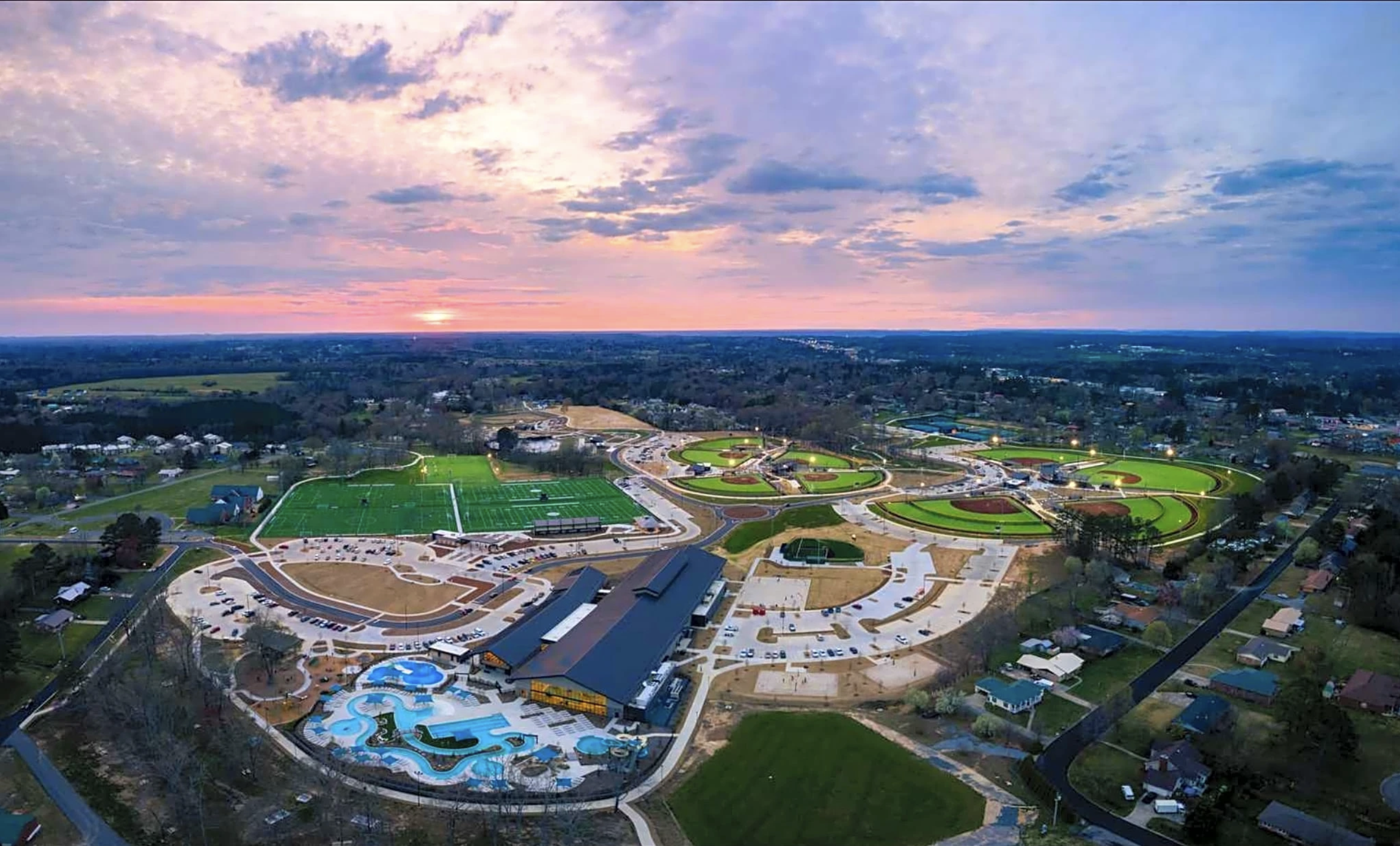 Aerial view of a large recreational facility at sunset, featuring sports fields, a waterpark with slides, parking lots, and surrounding residential area.